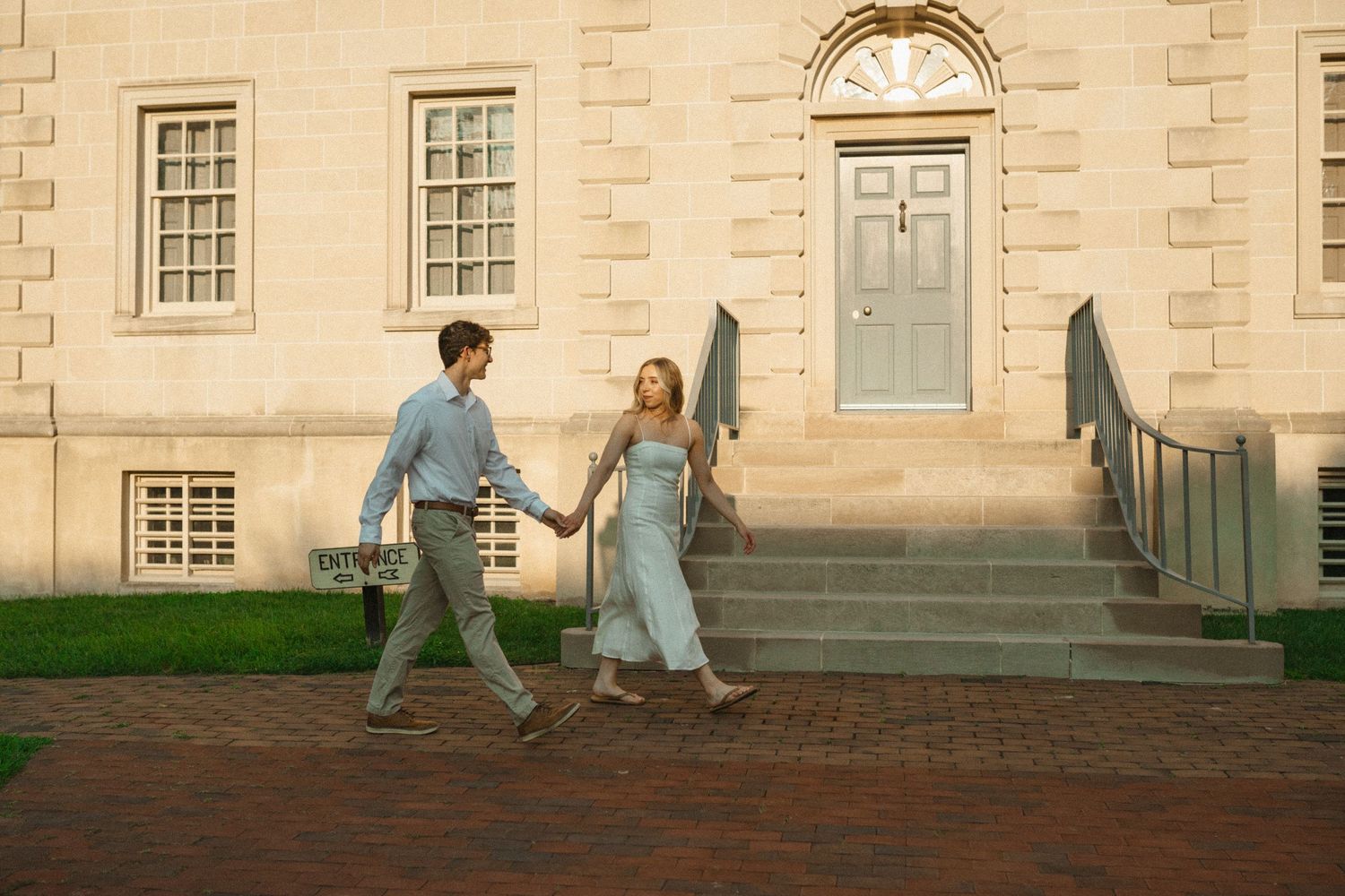 A couple holds hands and walks across a brick path in front of a historic stone building with a green door during an engagement session.