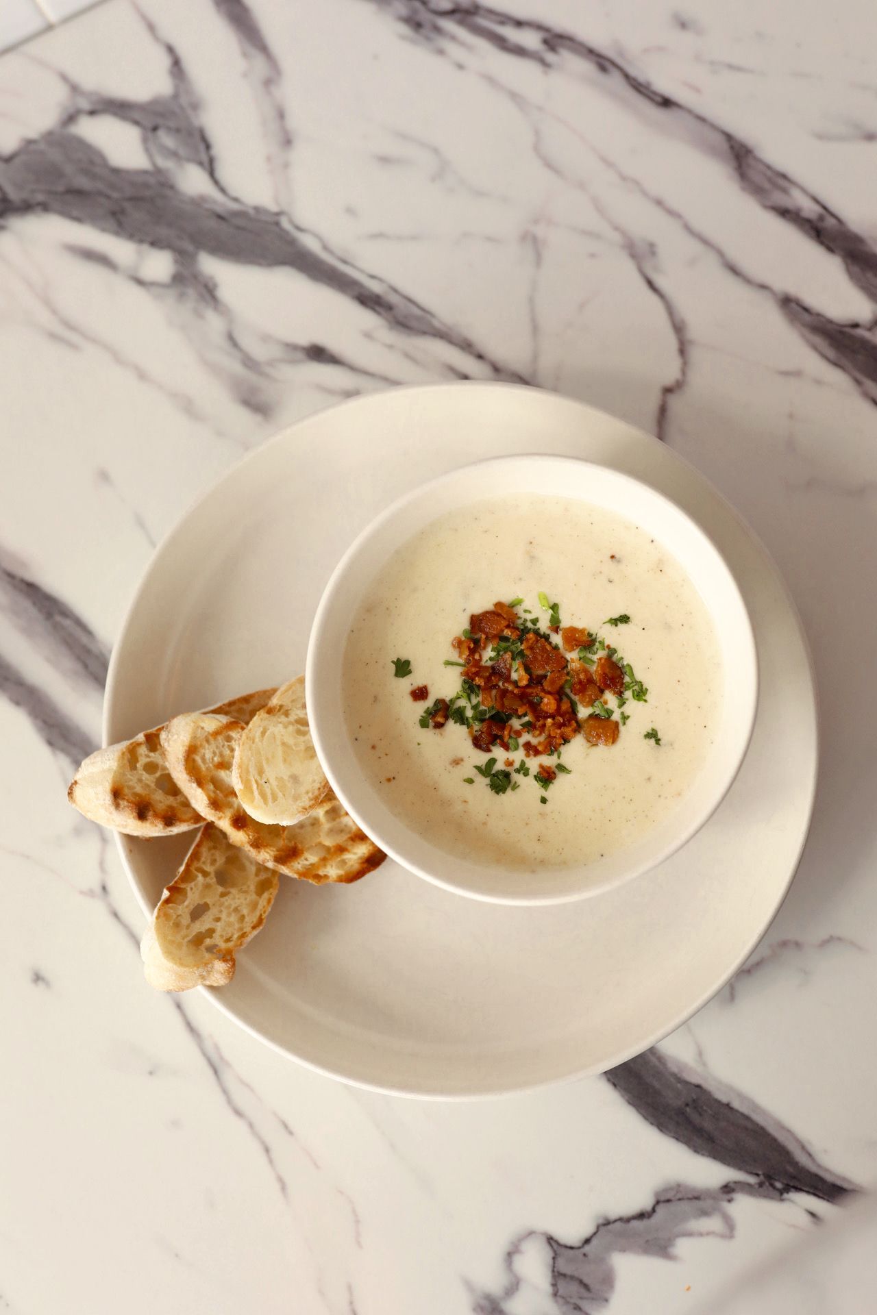 Overhead view of creamy soup topped with crispy bacon and herbs in a white bowl with toasted bread slices on a marble table.