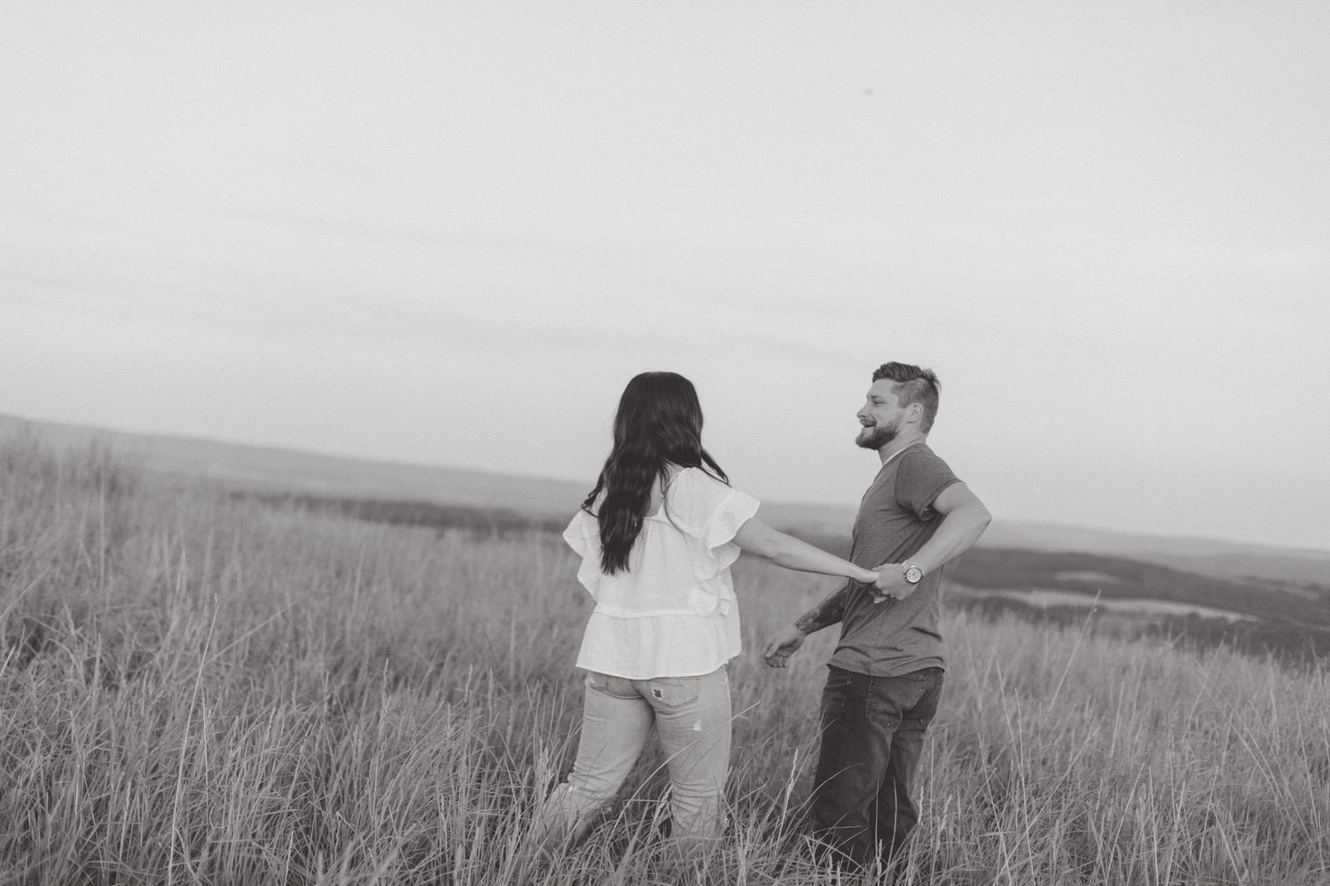Black and white engagement photo of a couple holding hands and laughing in tall grass on a hilltop overlooking the countryside.