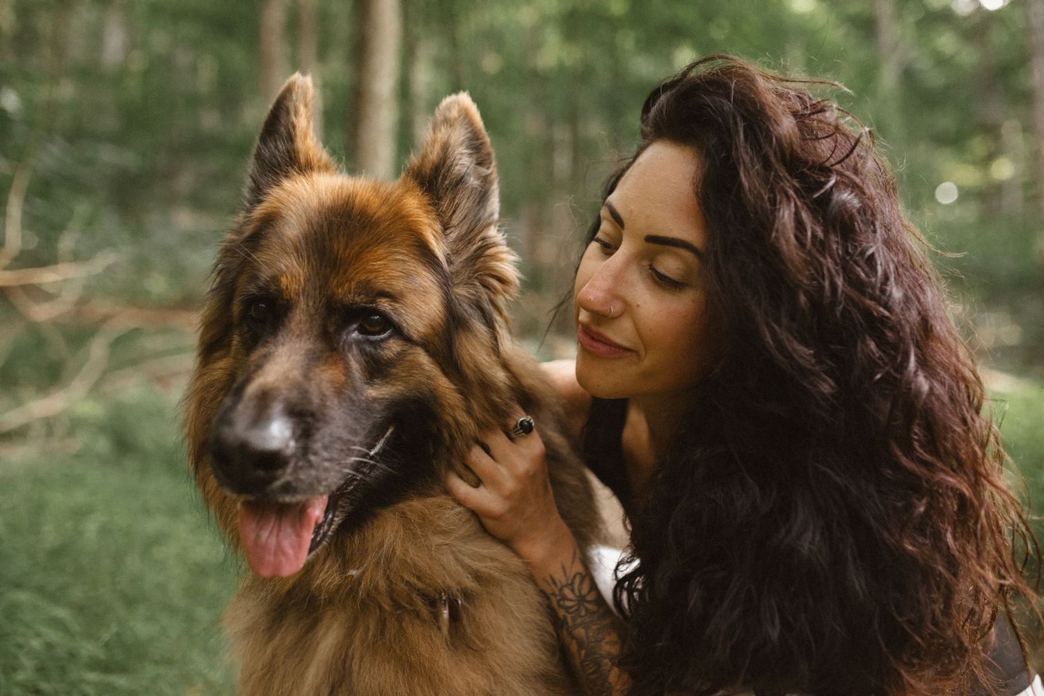 Close-up of a smiling woman with long curly hair embracing her German Shepherd dog in a green forest.