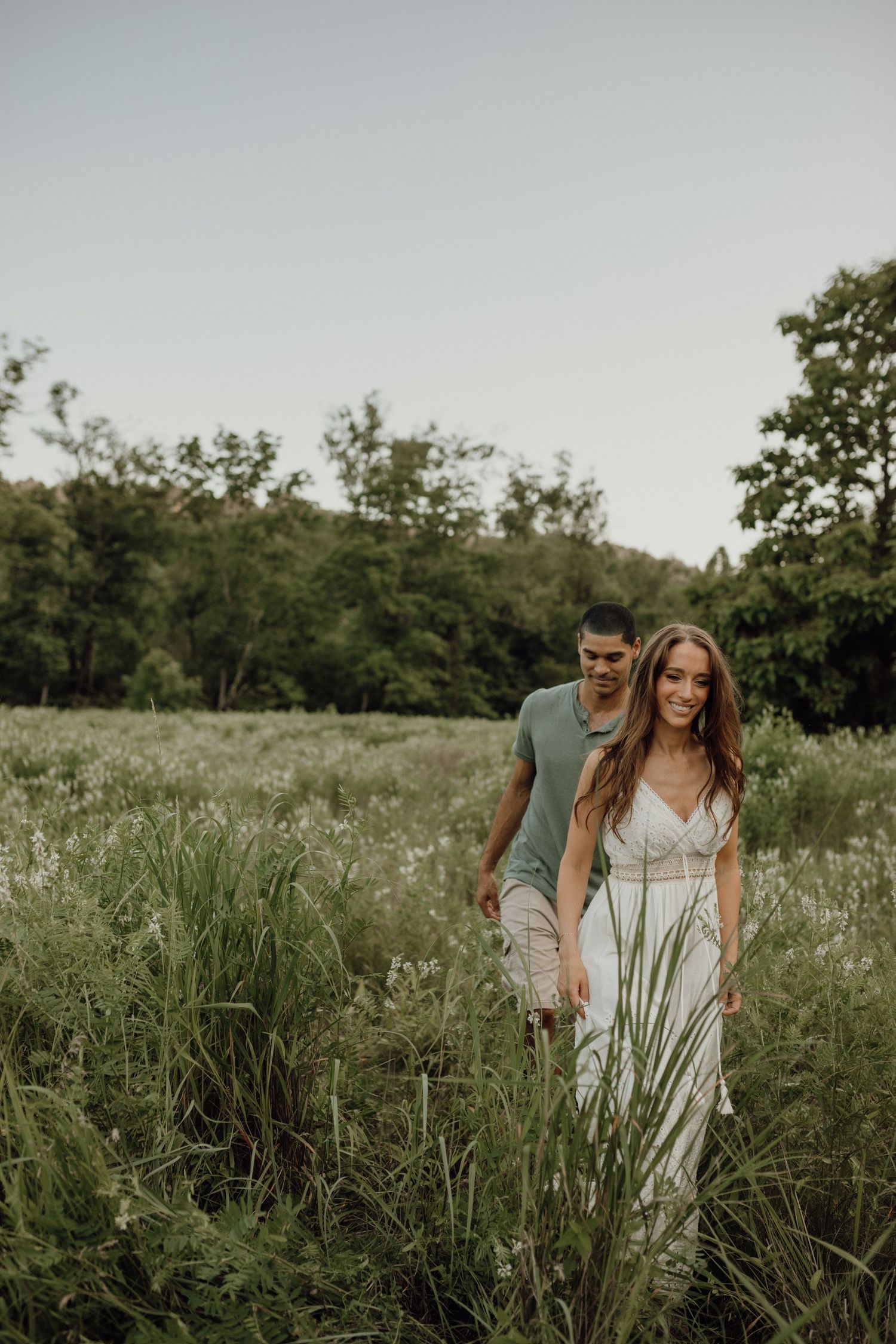 A smiling woman in a white dress walks through tall meadow grass with a man in a green shirt behind her.