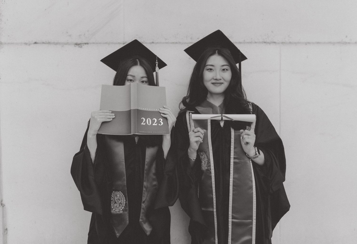 Two graduates in caps and gowns pose in black and white, one holding a 2023 book over her face, the other holding a frame.