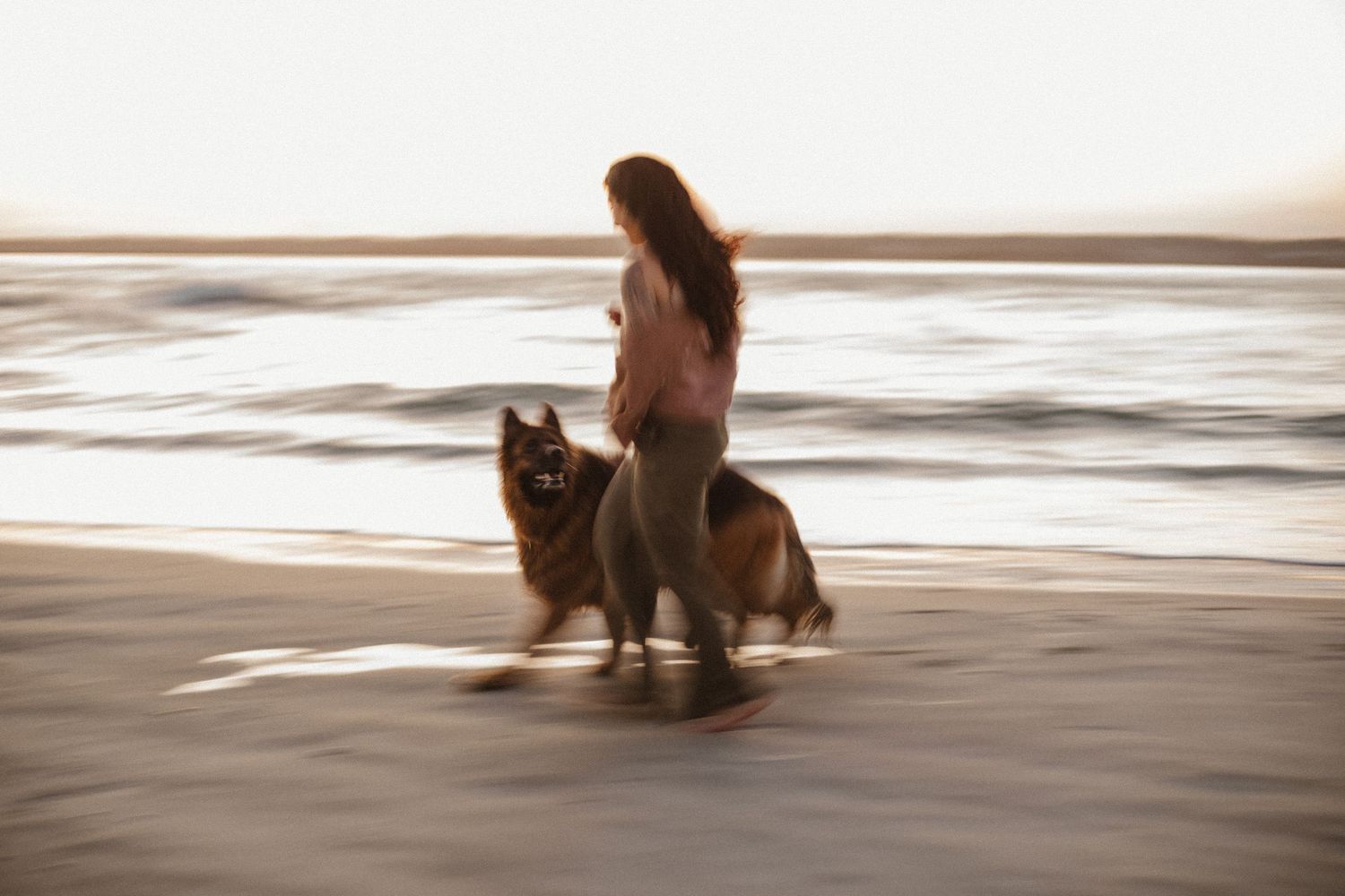 Motion-blurred image of a woman walking with her German Shepherd dog along the wet sand by the sea.