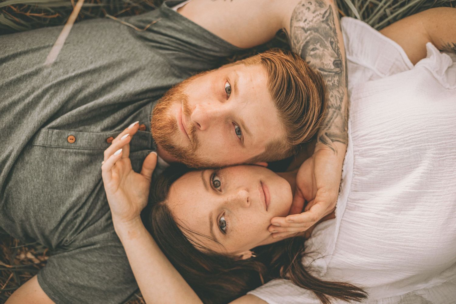 Overhead view of a bearded man and a woman lying close together in grass, gazing up at the camera.