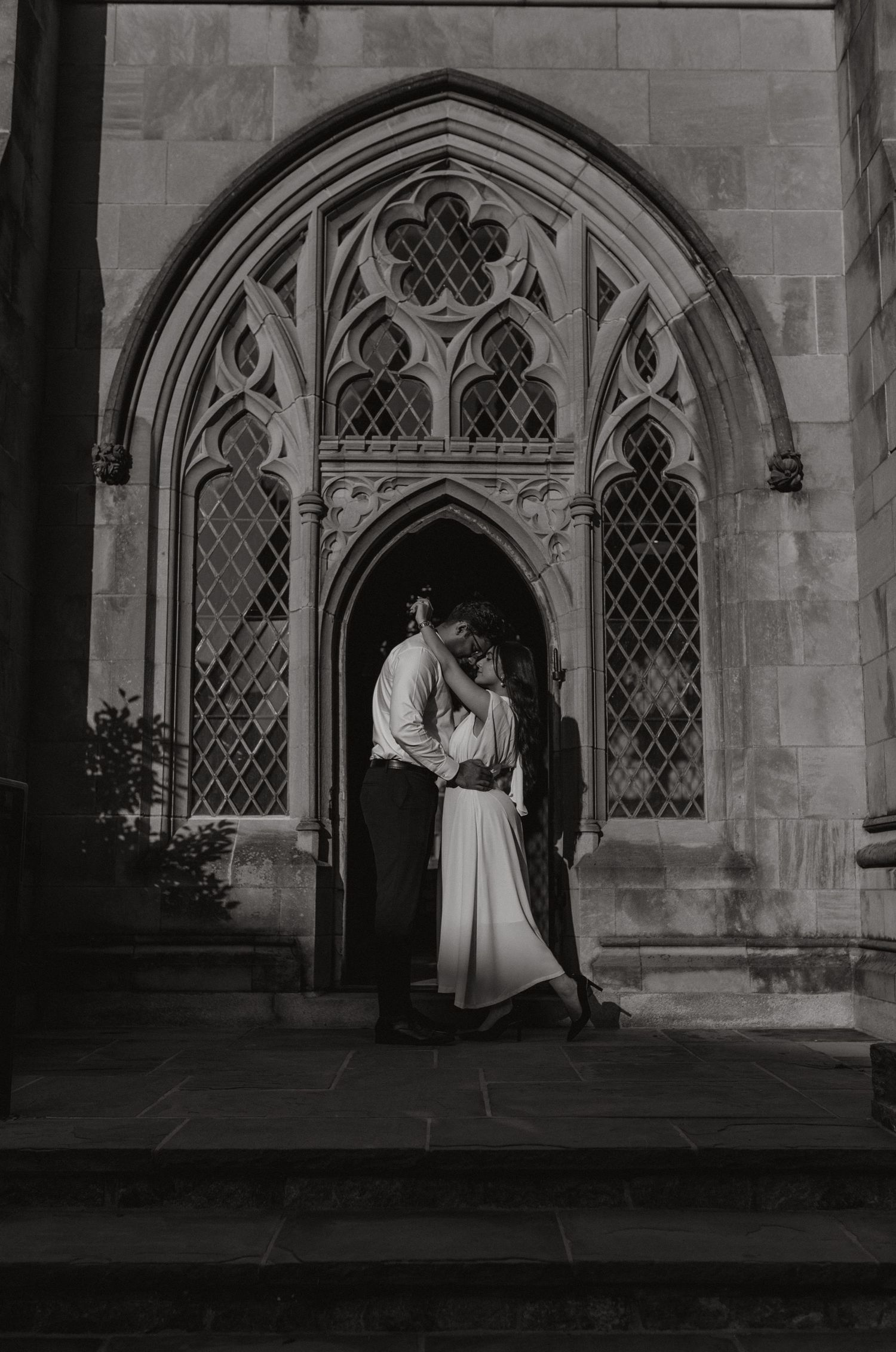 Black and white photo of a couple embracing in a Gothic stone doorway with tracery windows.