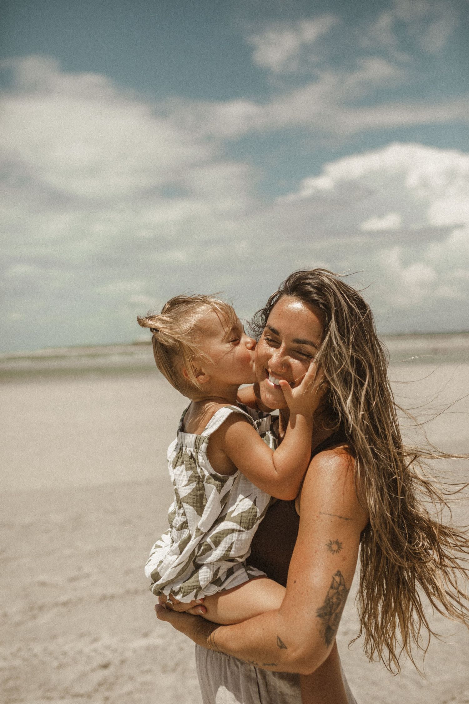 Smiling mother holding her toddler who kisses her cheek on a sandy beach under a bright cloudy sky.