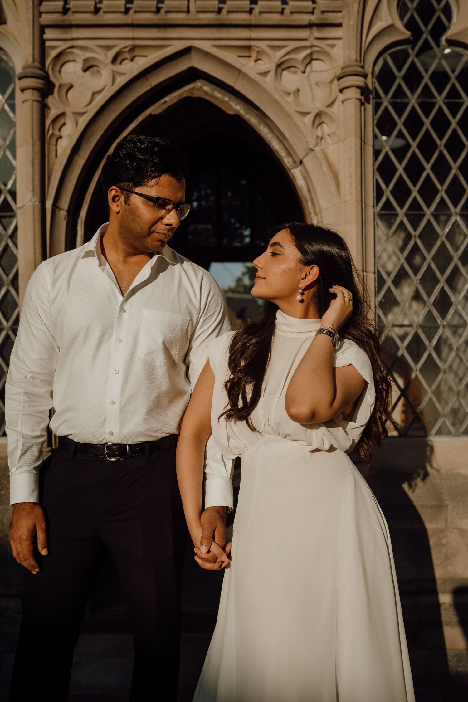 Smiling couple hold hands and gaze at each other in front of a Gothic stone building in warm sunlight.