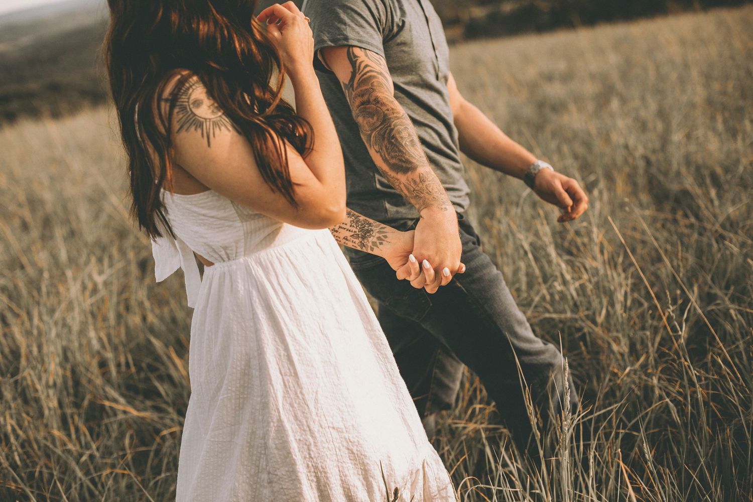 A woman in a white dress and a tattooed man walk hand in hand through a golden grass field, seen from behind.