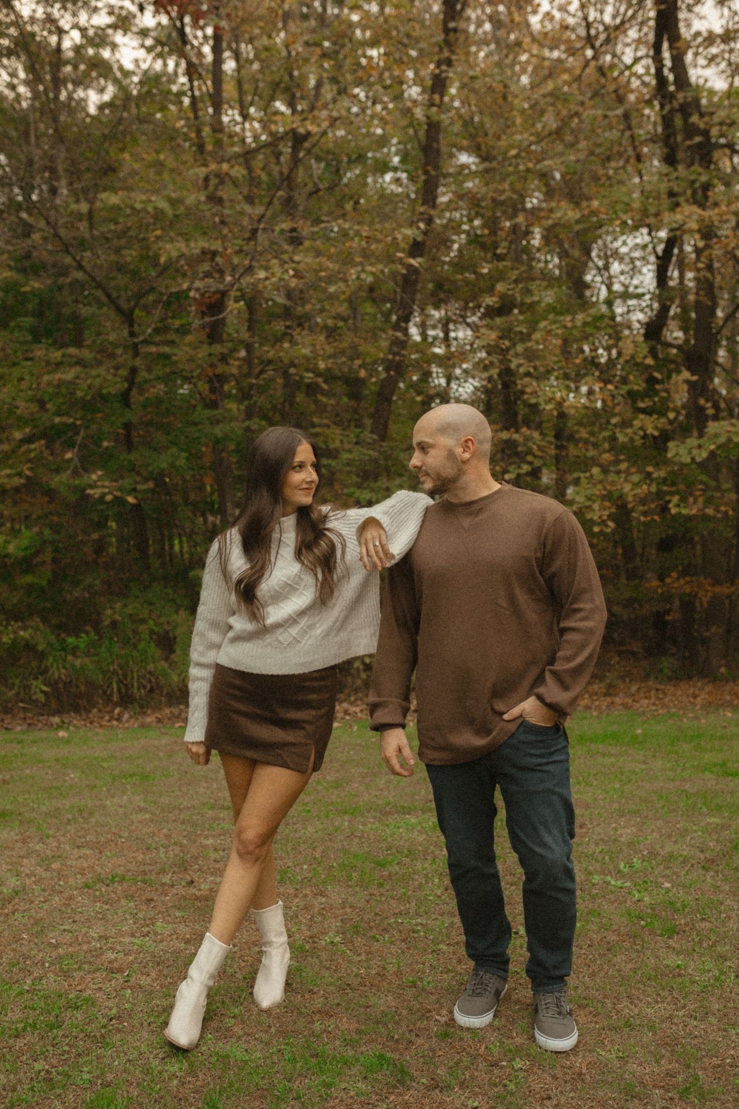 Couple posing casually in an autumn woodland clearing, the woman leaning on her partner's shoulder in a sweater and boots.