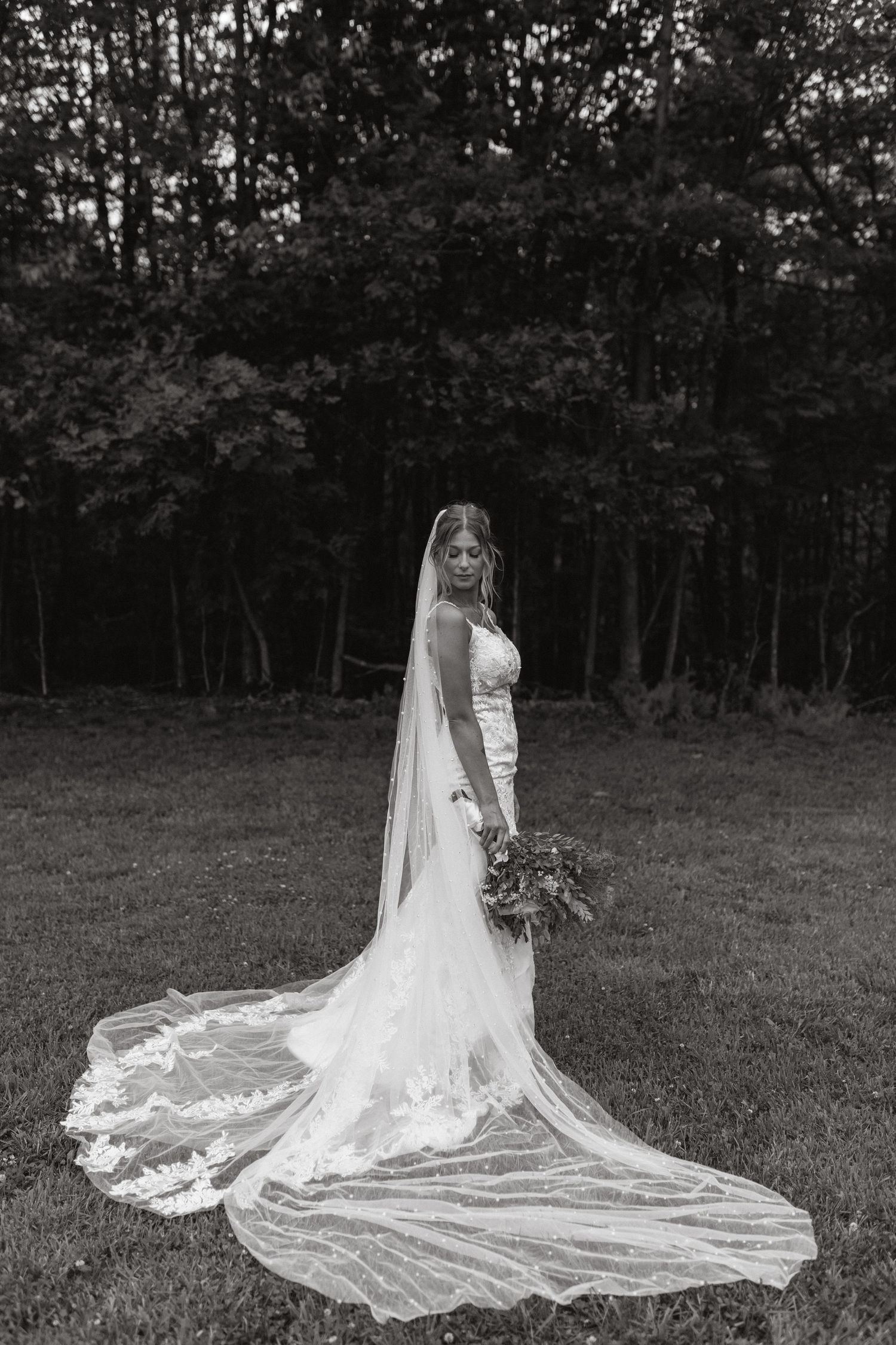Black and white portrait of a bride in a lace gown with a long veil holding a bouquet in a forest clearing.