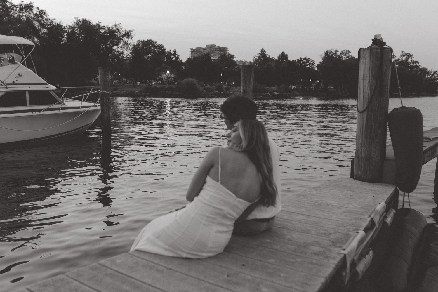 Black and white photo of a woman in a white dress sitting on a dock looking out at boats on the water.