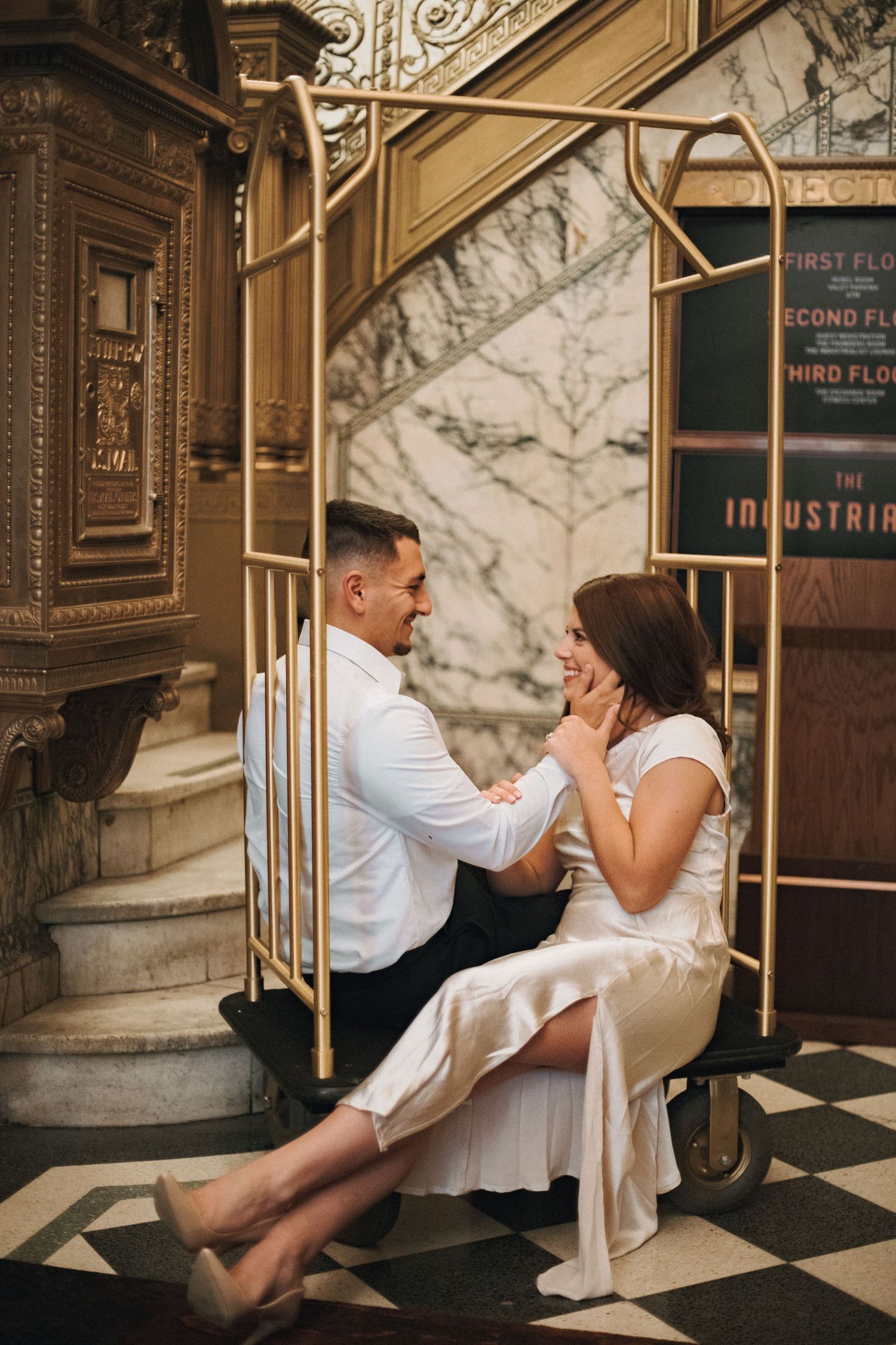 Smiling couple sitting together on a gold luggage cart in a grand vintage building with marble walls and checkered floors.