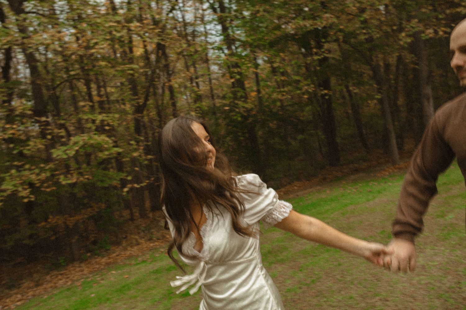 Woman in a white dress playfully leads a man by the hand along a grassy path lined with autumn trees.