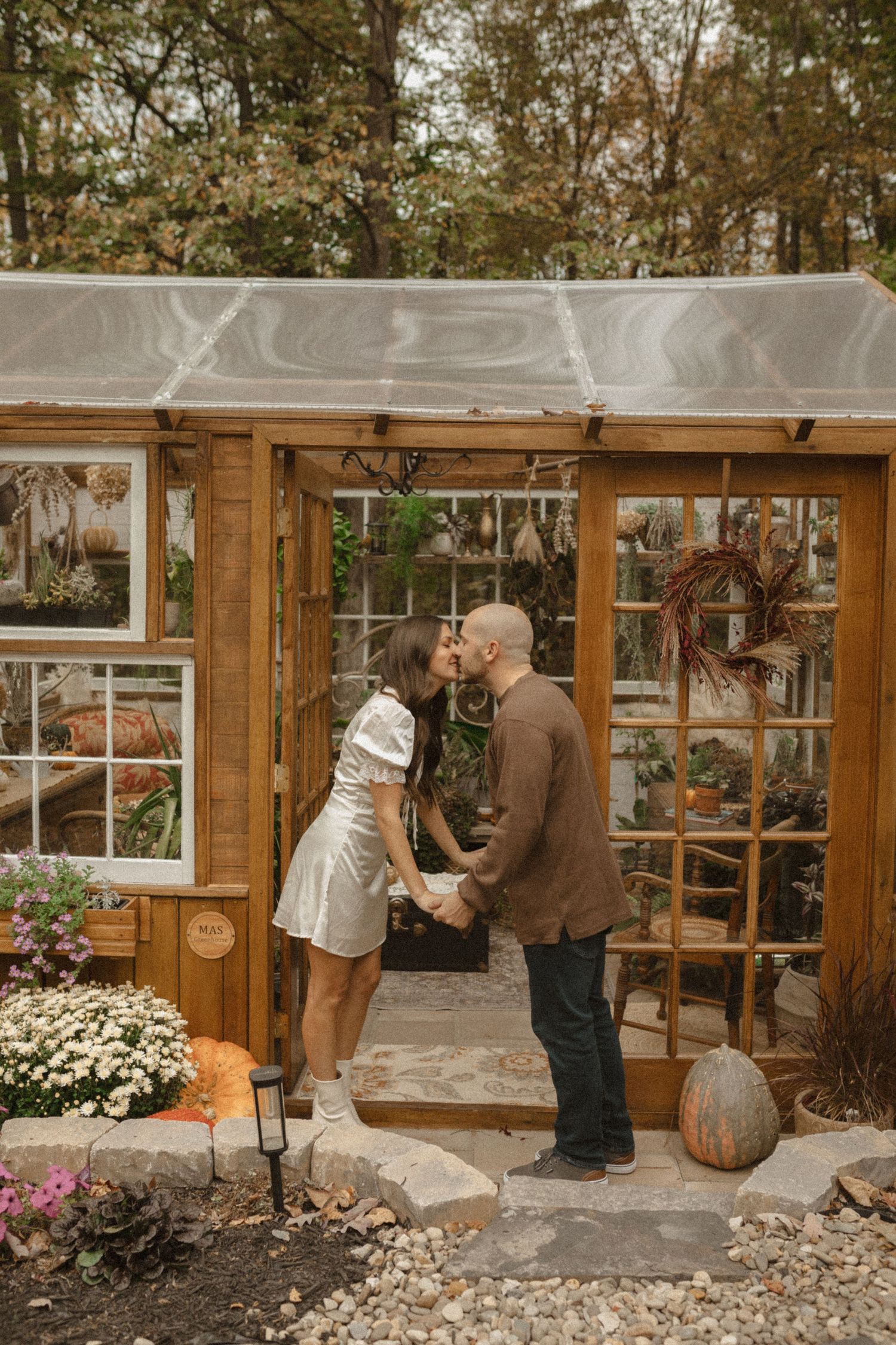 Couple shares a kiss at the doorway of a wooden greenhouse surrounded by autumn foliage, mums, and pumpkins.