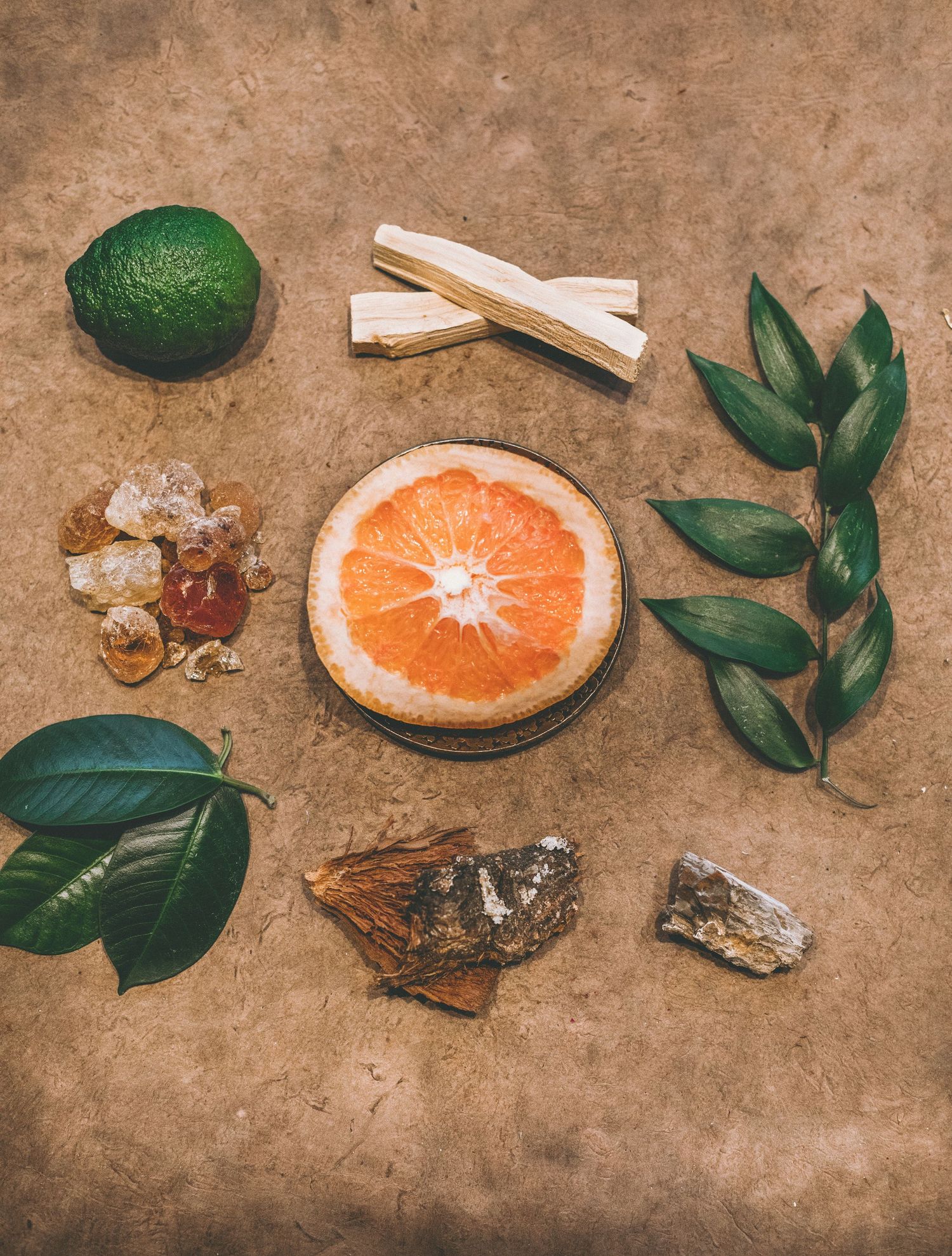 Flat lay of a sliced grapefruit, a lime, palo santo sticks, resin crystals, green leaves and a mineral on a tan surface.