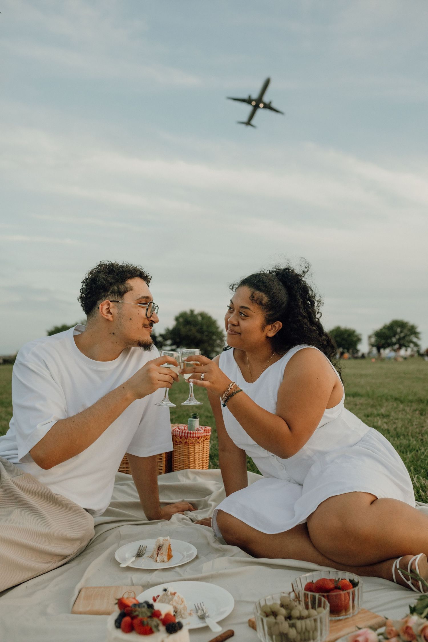 A couple clinks glasses at a picnic with fruit and desserts as an airplane flies across the sky above.