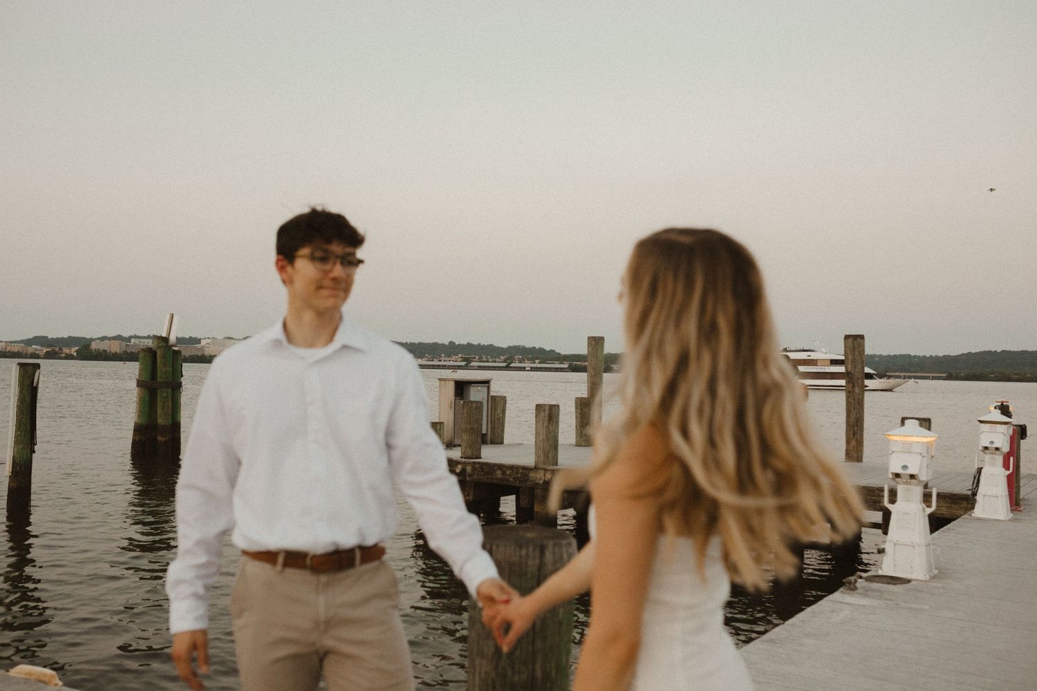 Man in a white shirt holds hands with a woman in a white dress on a wooden dock at sunset by the water.