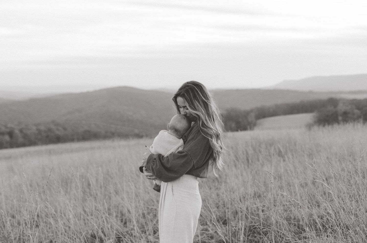Black and white photo of a woman with long hair holding a baby in a grassy field with rolling hills in the background.