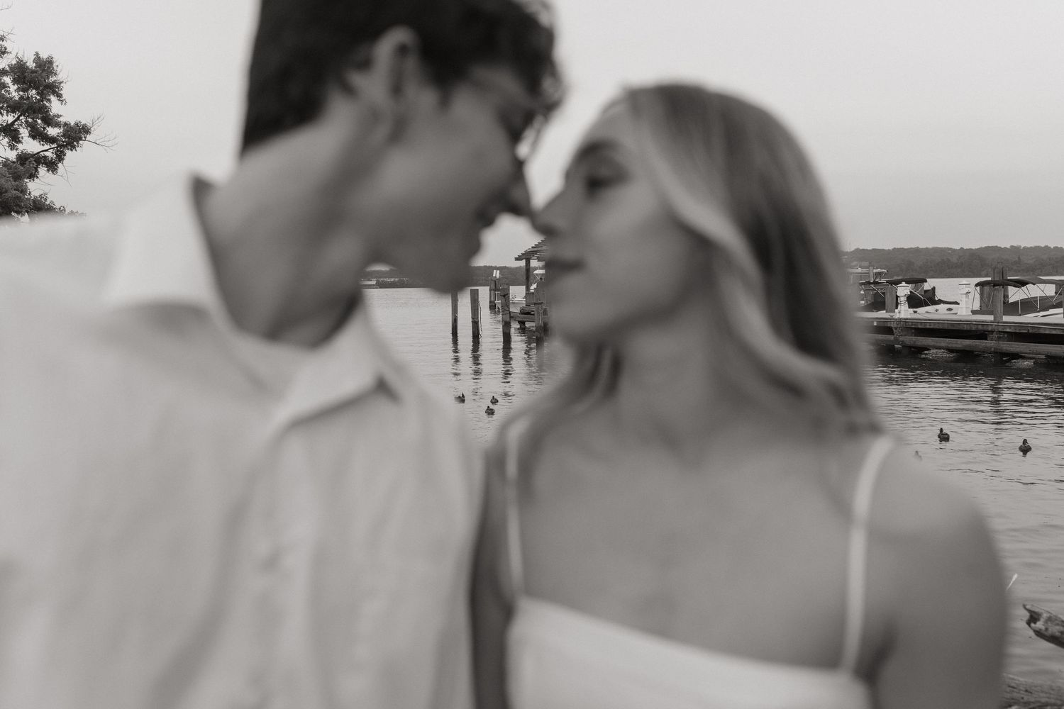 Blurry black and white close-up of a couple about to kiss in front of a calm waterfront with boats.