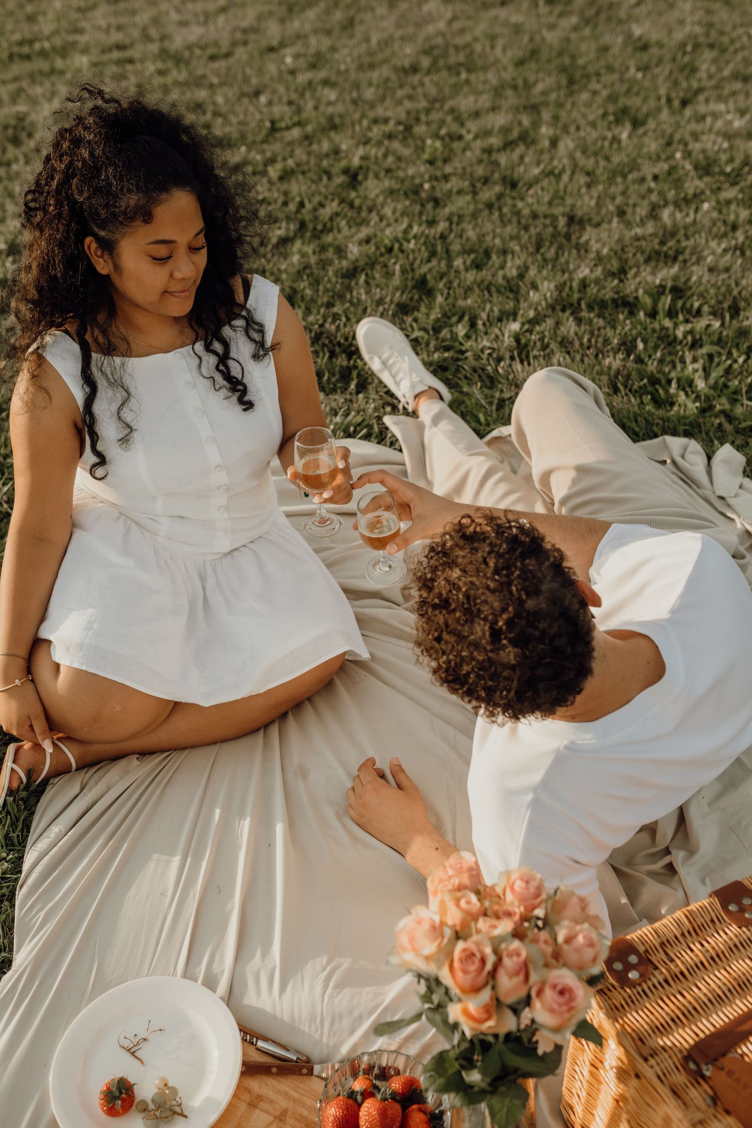 Overhead view of a couple in white clothing toasting drinks on a picnic blanket in a grassy park.