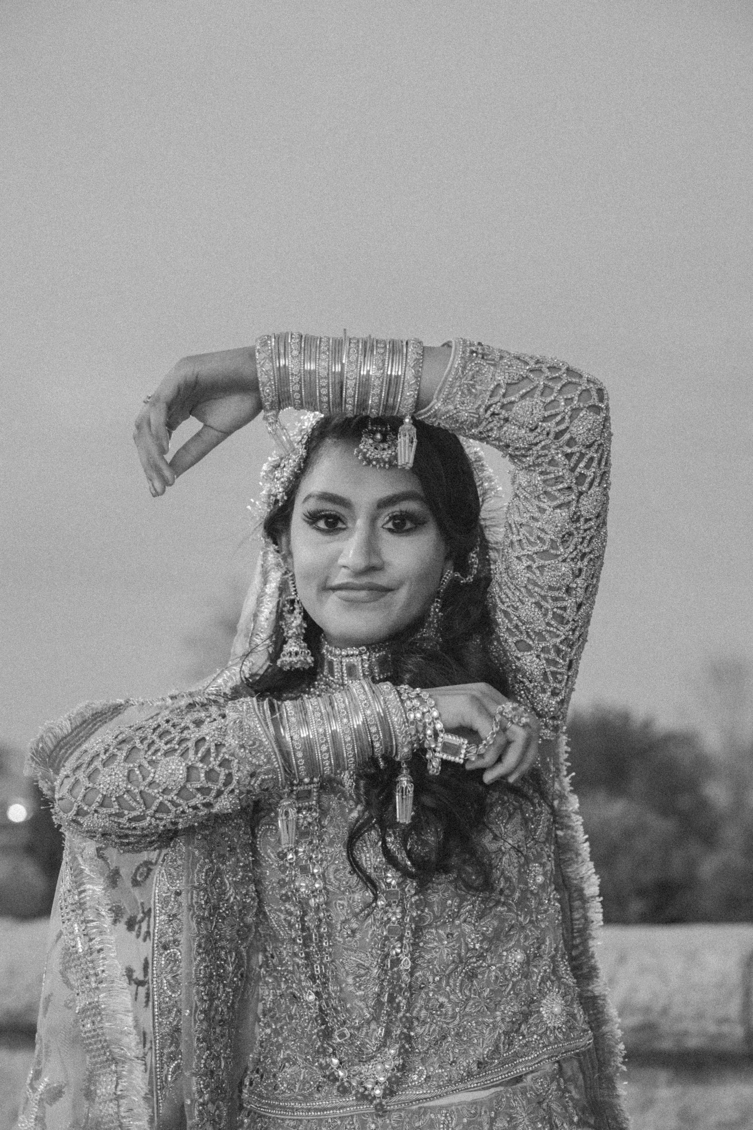 Black and white portrait of a bride in ornate embroidered attire and bangles posing with arms raised overhead.