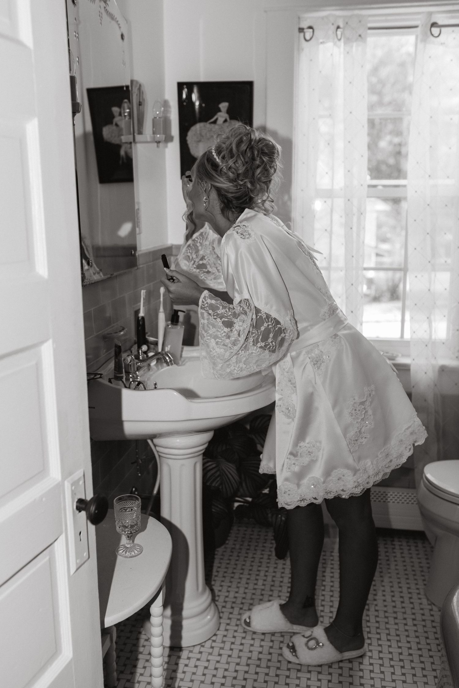Black and white photo of a bride in a lace robe and slippers leaning over a pedestal sink to get ready in a bright bathroom.