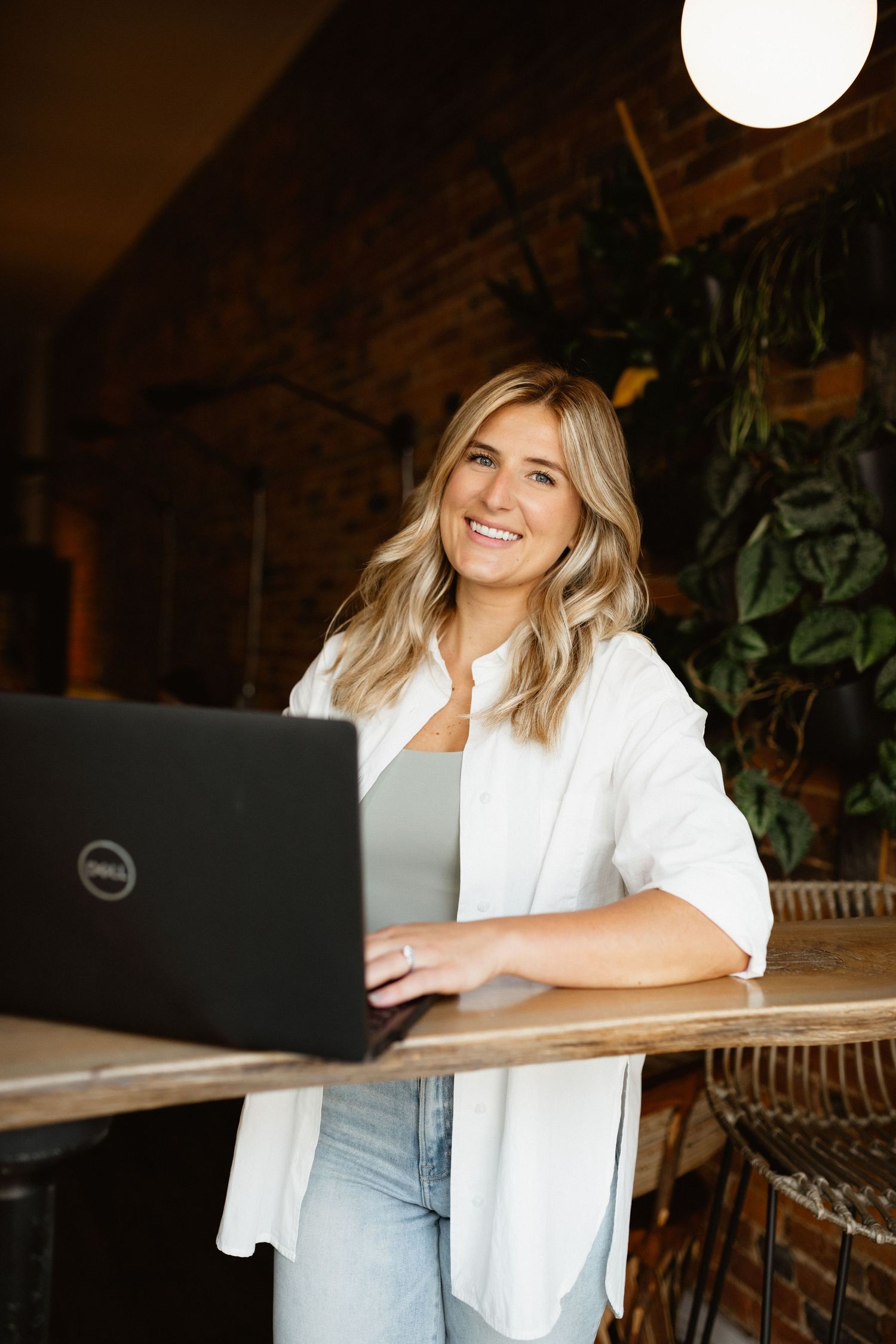Smiling blonde woman in a white shirt leans on a wooden table with a Dell laptop in a cozy brick café.