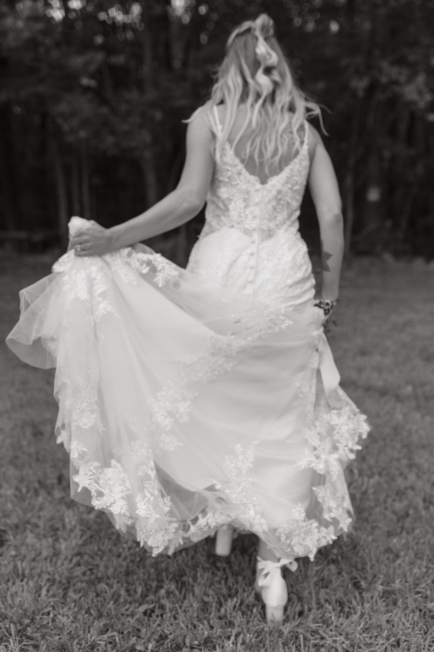 Black and white photo of a bride in a lace gown holding her flowing skirt as she walks across the grass.