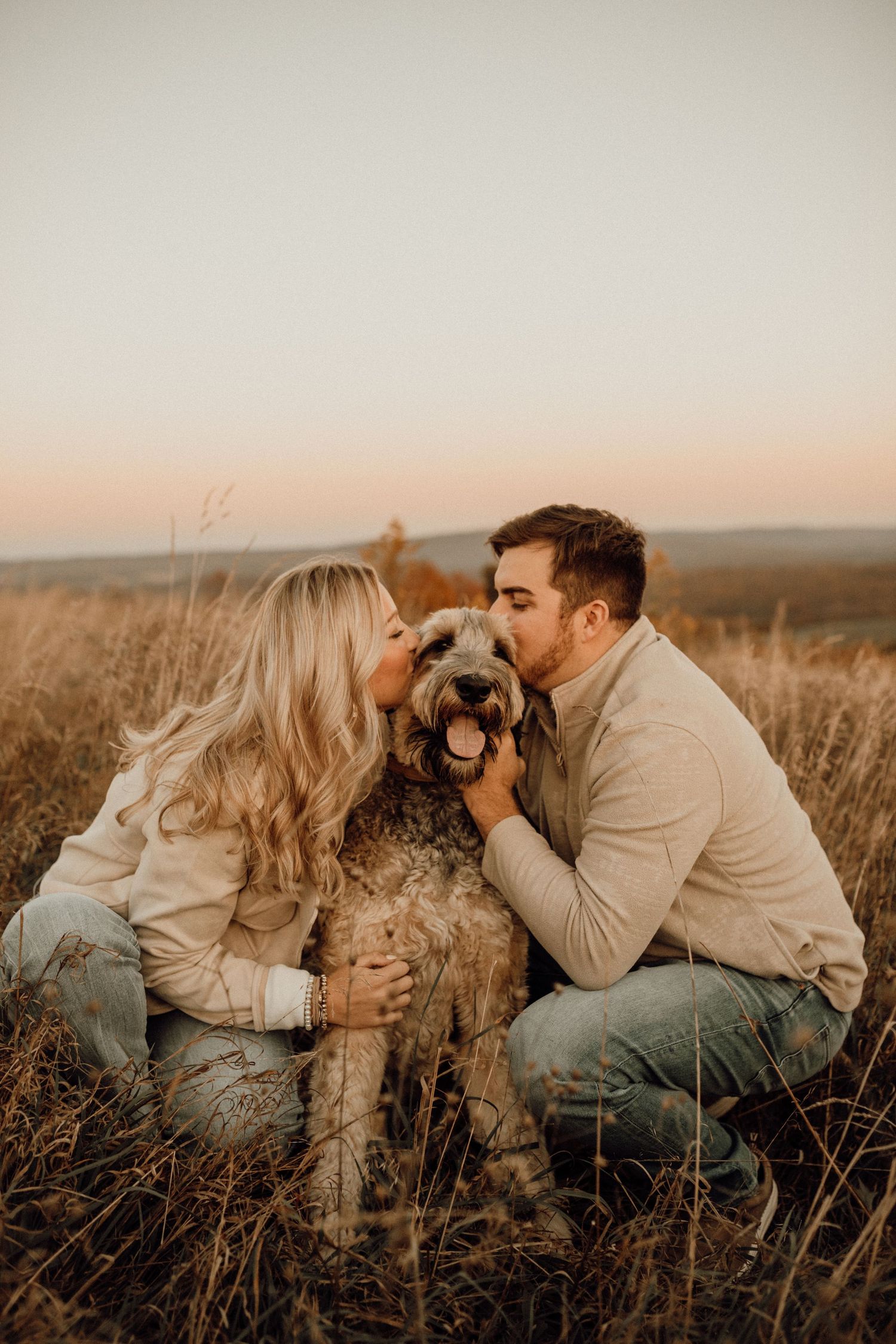 Couple kneeling in tall autumn grass kissing their shaggy dog on the head under a soft pastel sunset sky.