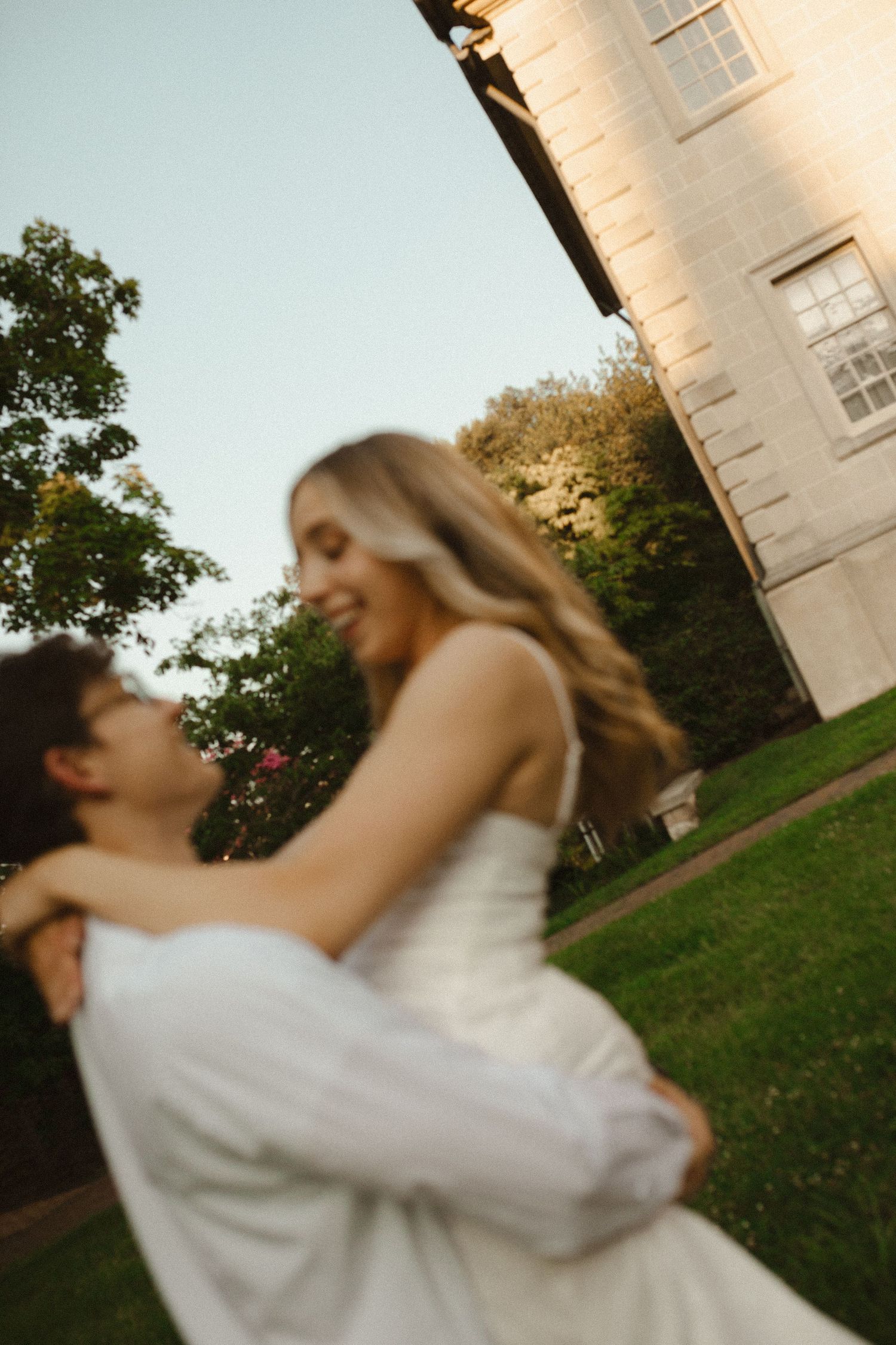 Smiling man lifts a woman in a white dress in a joyful embrace on a green lawn beside a historic building.