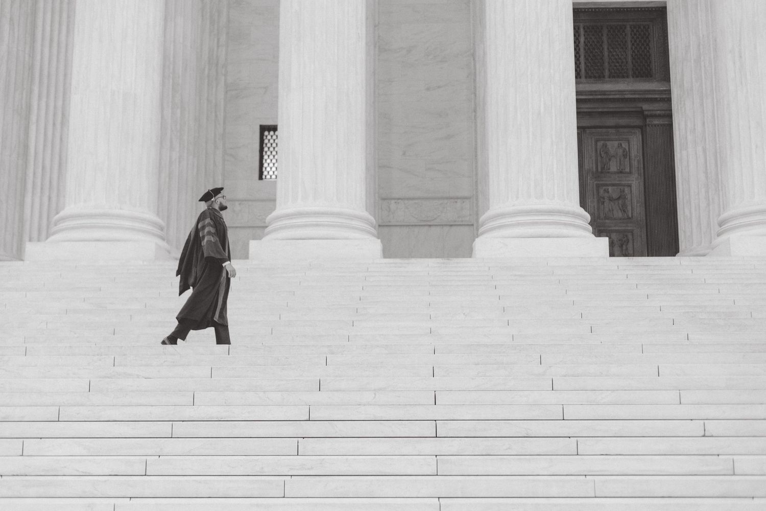 Black and white image of a person in graduation gown walking up wide steps toward a columned courthouse building.