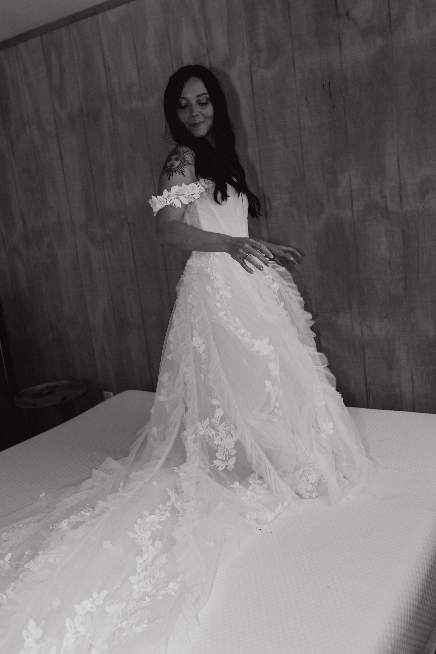 Black and white photo of a dark-haired bride in an off-shoulder lace wedding dress with a long train standing on a bed.