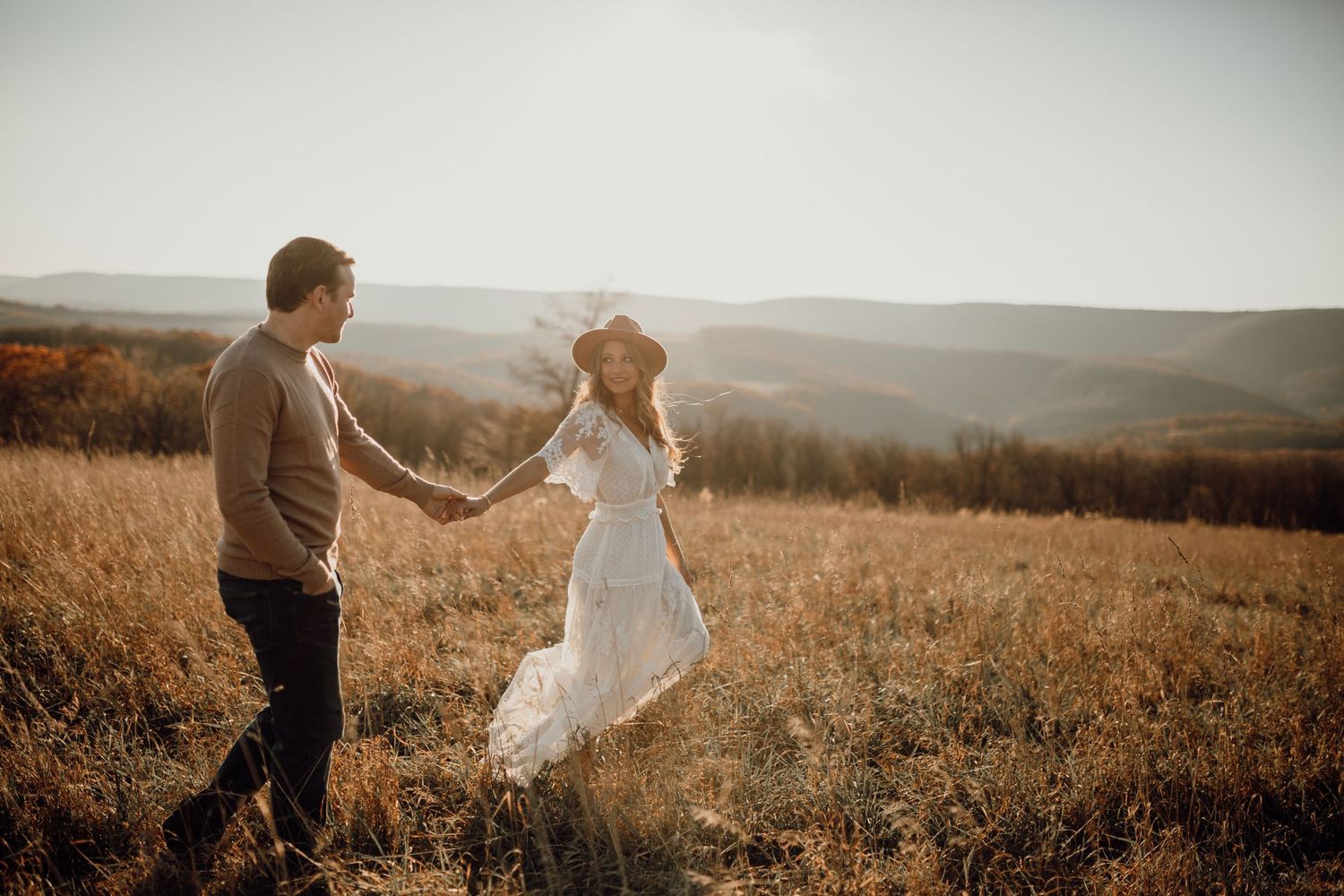 Man in a sweater leads a woman in a white dress and hat by the hand through a sunlit autumn meadow.