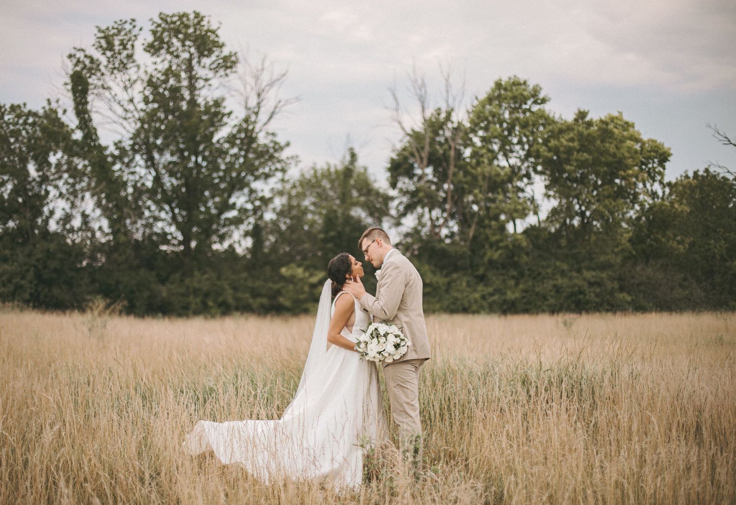 Bride in a white gown and groom in a tan suit embrace with a bouquet in a tall golden grass field with trees behind them.