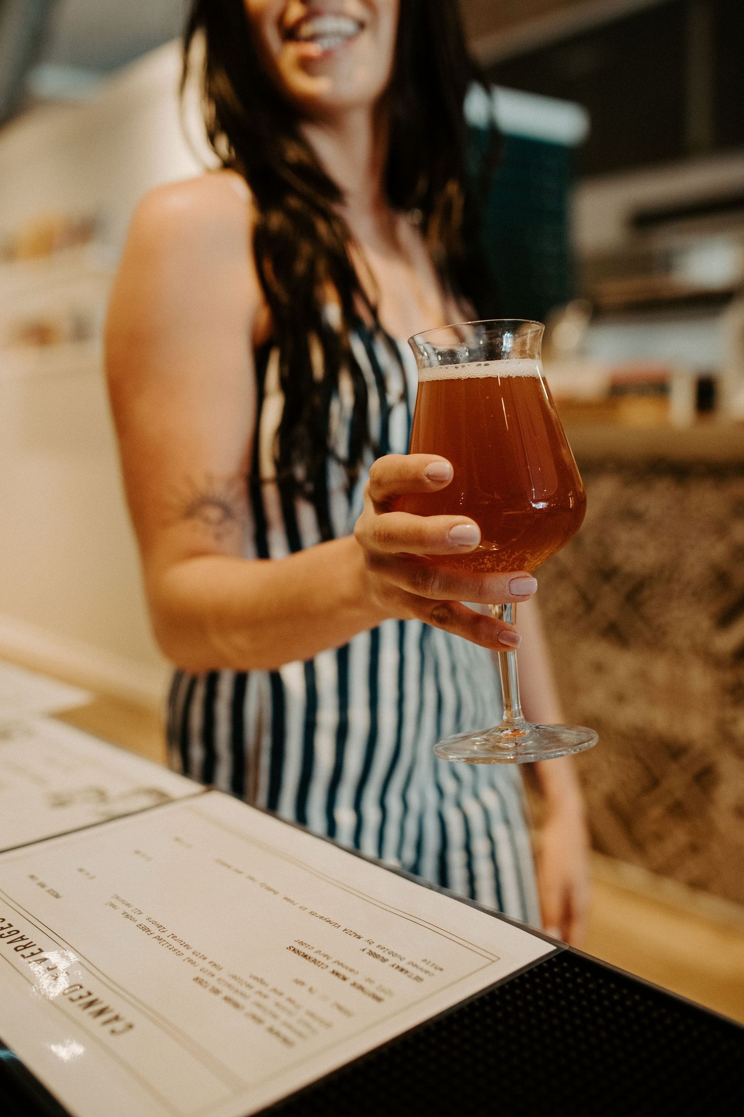 Smiling woman in a striped apron holds a glass of amber craft beer at a bar beside a menu.