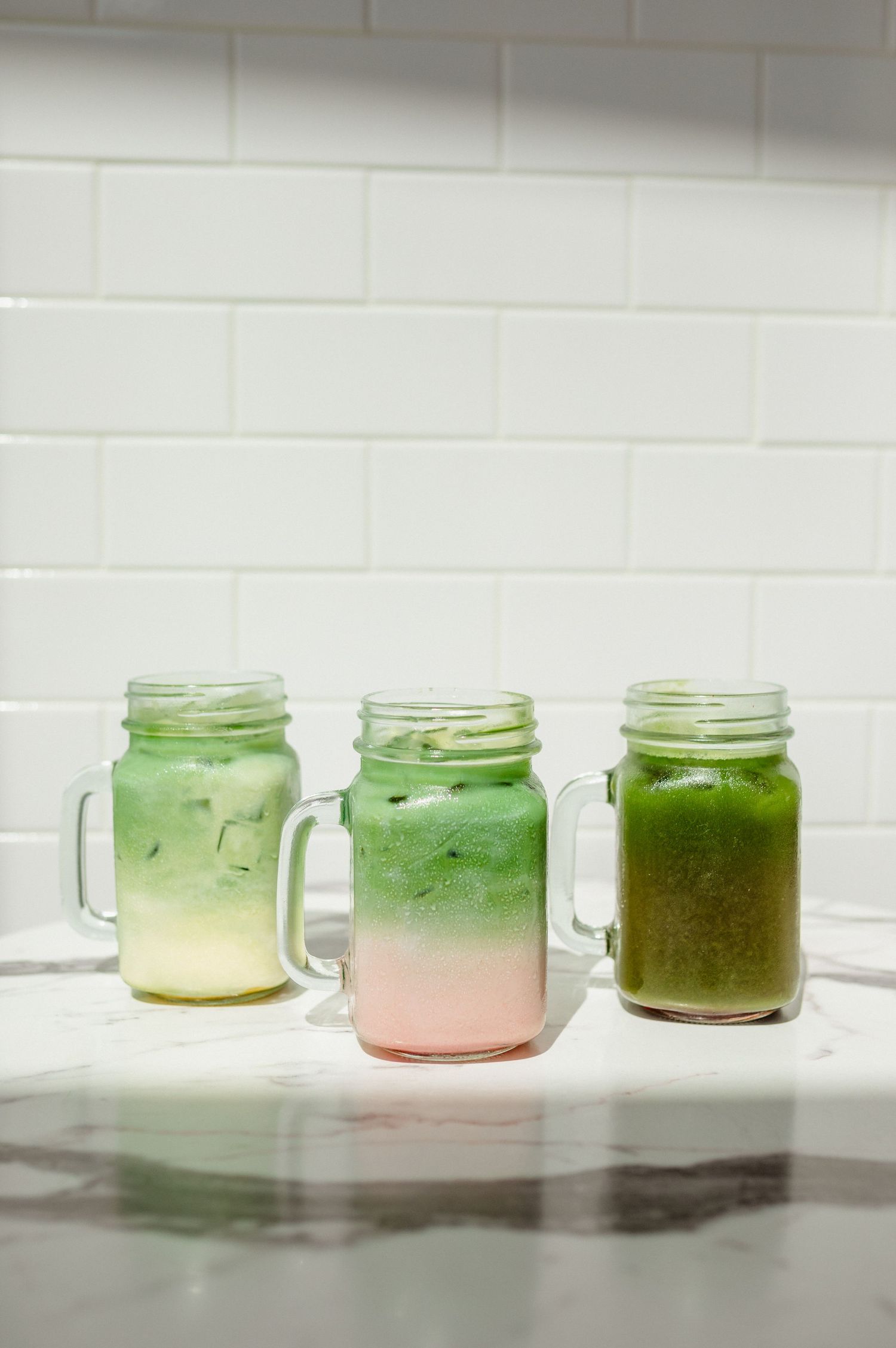 Three layered iced matcha and fruit drinks in glass mason jars lined up on a marble counter by a white tile wall.