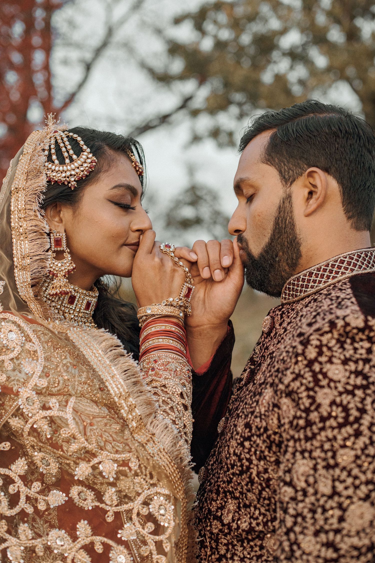 Bride in ornate gold embroidered South Asian wedding attire gently kisses her groom's hand as he wears a rich maroon embroidered sherwani.