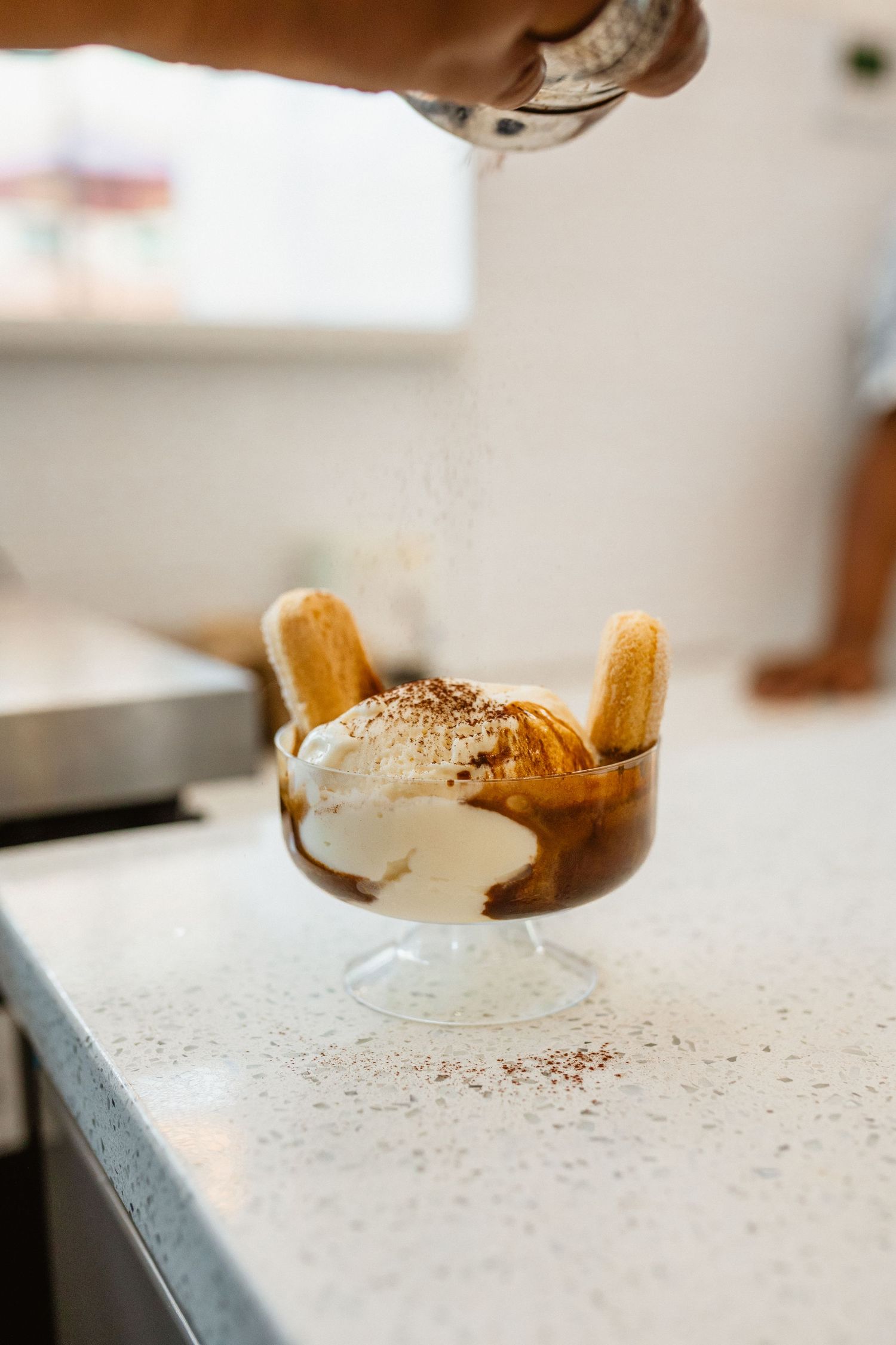 Dessert glass with vanilla ice cream, espresso and ladyfingers being dusted with cocoa powder at a counter.