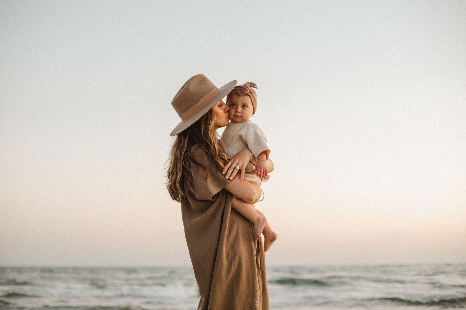 Mother in a wide-brim hat and tan wrap kisses her baby on the beach at sunset with ocean waves behind.