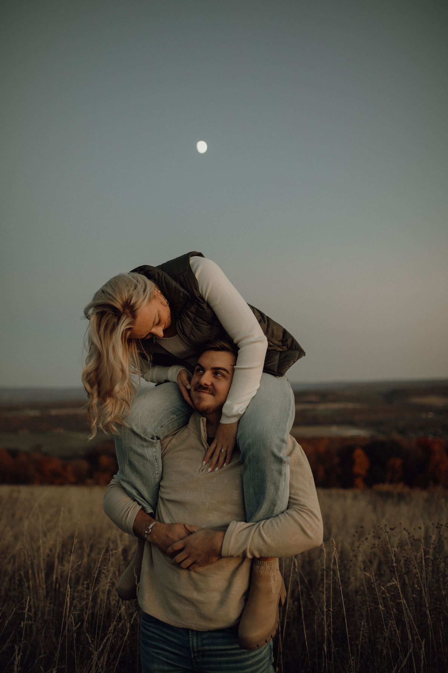 A playful woman sits on a smiling man's shoulders in an open field at twilight, with autumn foliage and the moon above.