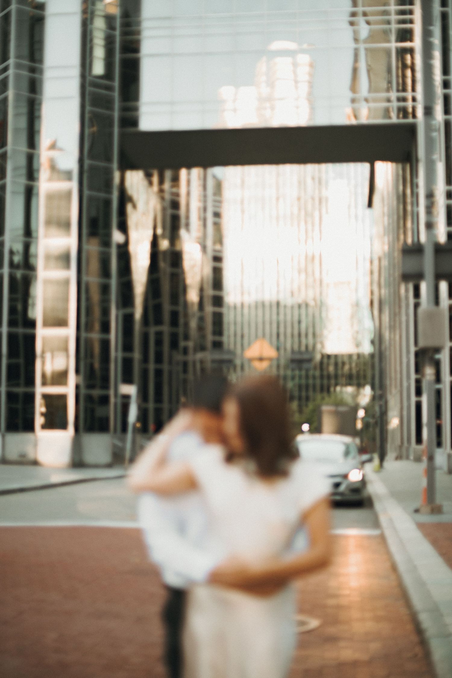 Blurred couple embracing on a brick city street with sunlit glass buildings and a pedestrian bridge behind them.