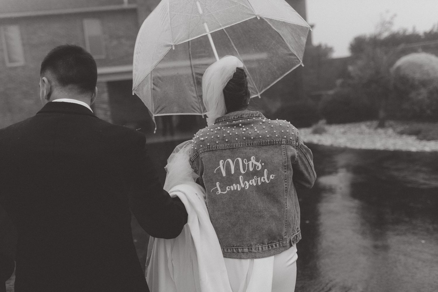 Black and white photo of a bride in a denim jacket reading Mrs. under a clear umbrella, walking with her groom in the rain.