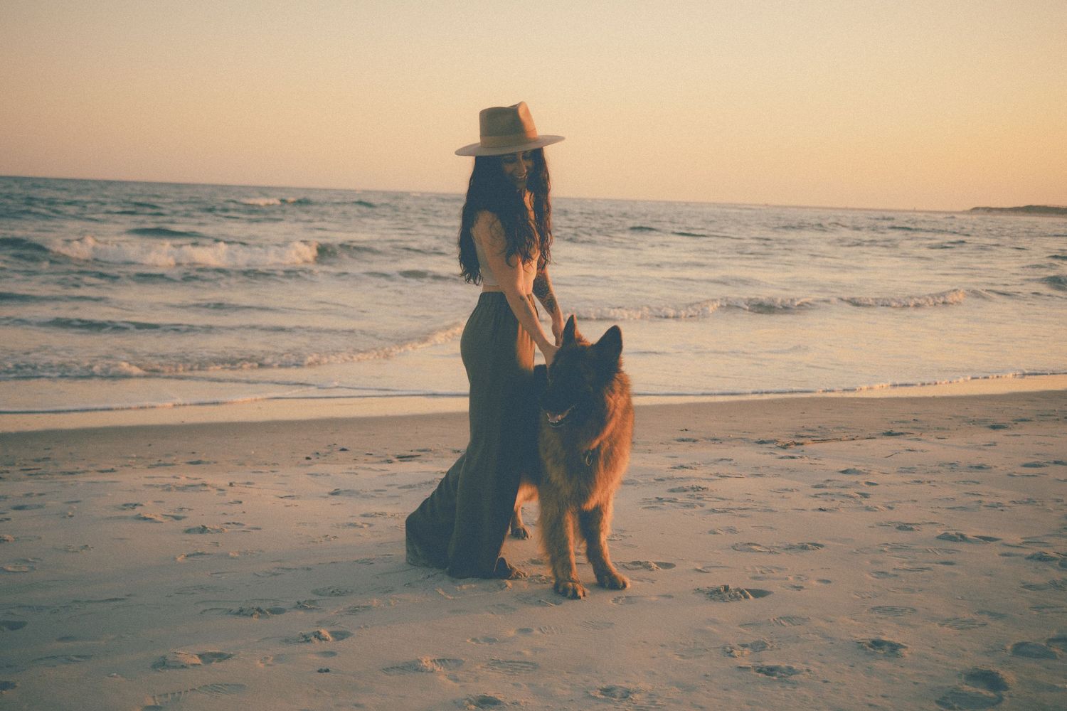 Woman in a wide-brim hat stands on a sandy beach at sunset with her German Shepherd dog beside her.