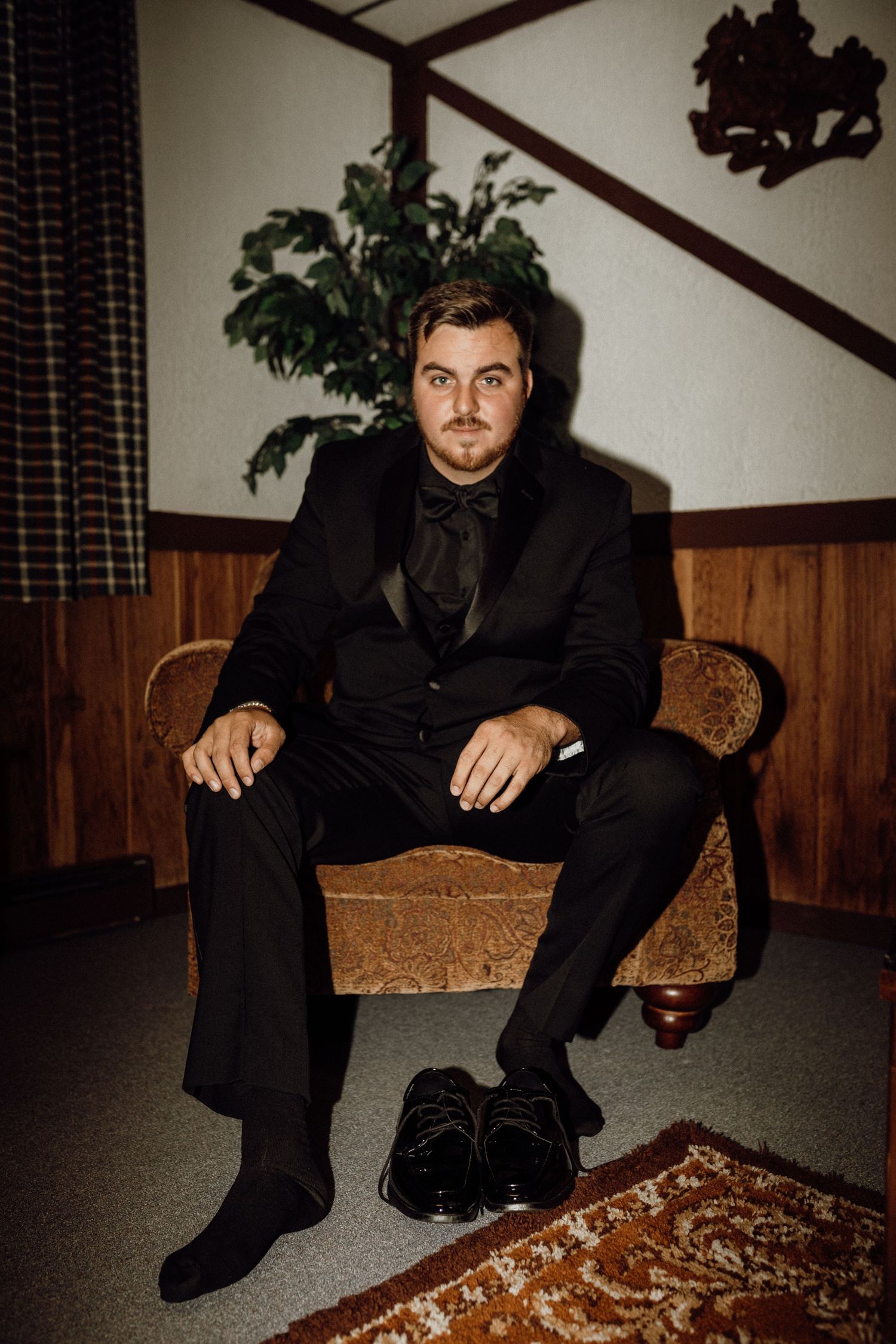 Groom in a black tuxedo and bow tie sits in a vintage armchair in a wood-paneled room with his shoes beside him.