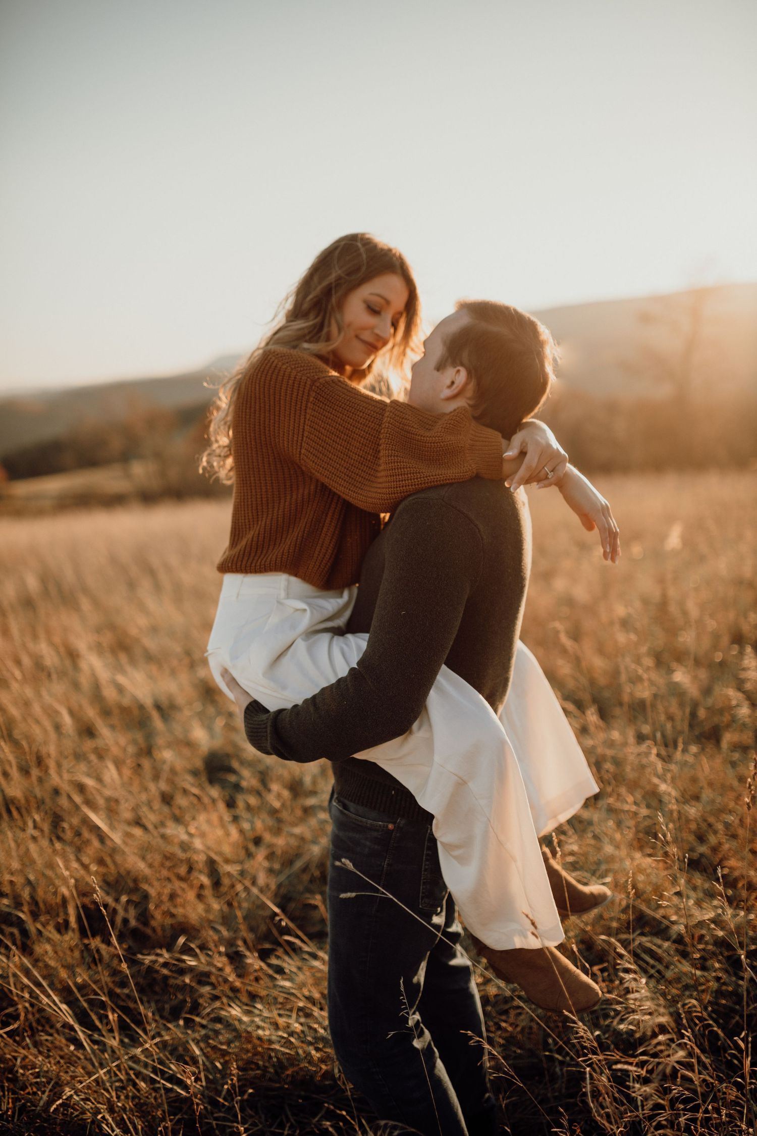 Man lifting his smiling fiancée in a brown sweater and white skirt in a golden field at sunset.