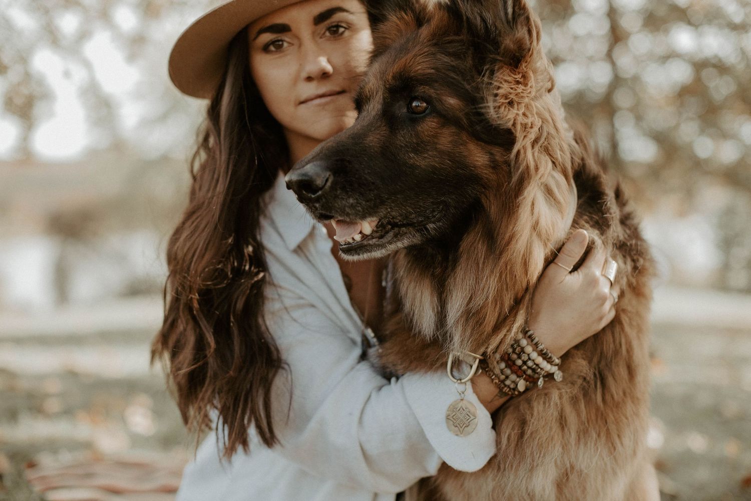 Woman in a hat and light jacket hugging a large German shepherd dog among autumn trees.