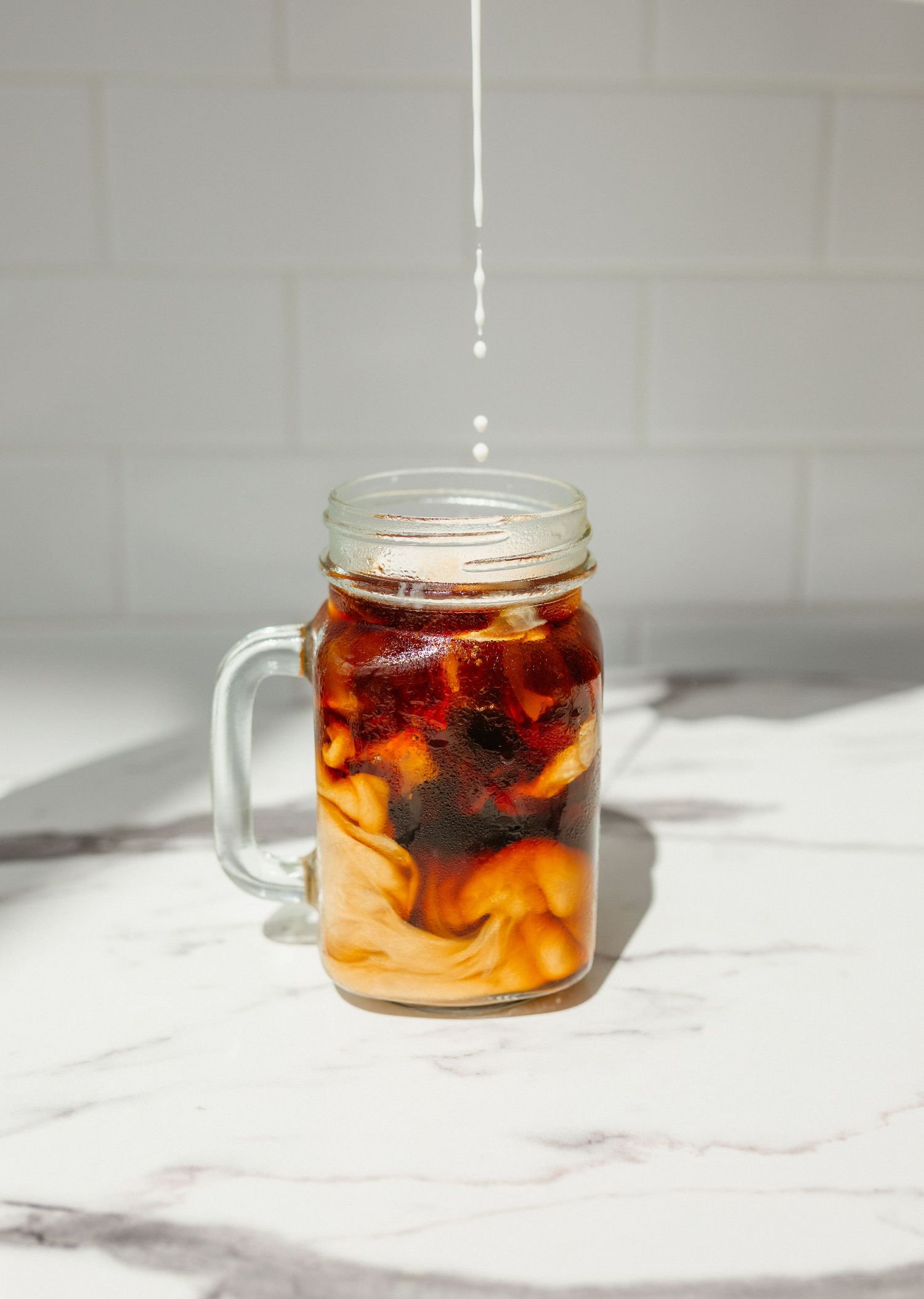 Iced coffee with cream swirling in a glass mason jar mug as milk pours in from above on a marble counter.