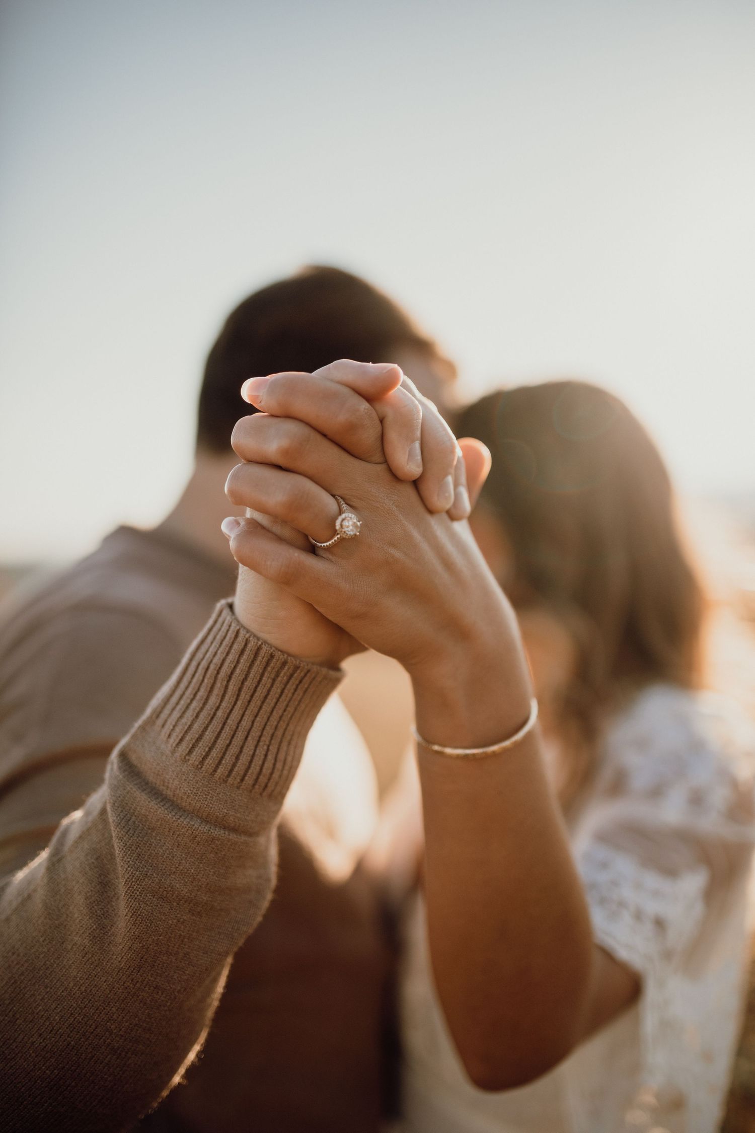 Couple clasp hands in the foreground to show off a diamond engagement ring with a softly blurred kiss behind.