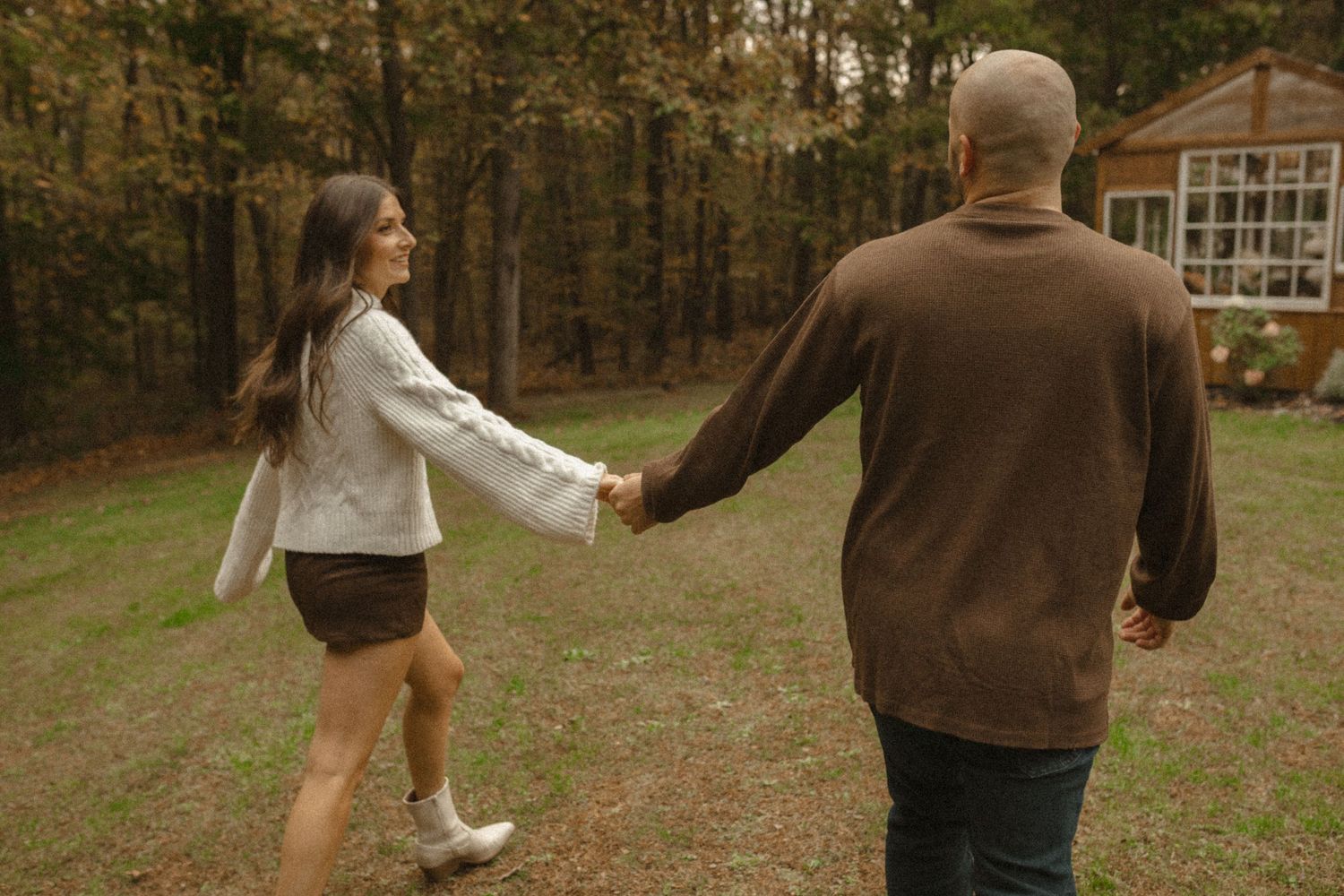 Woman in a white sweater leading her partner by the hand across a grassy field toward a small cabin in autumn.