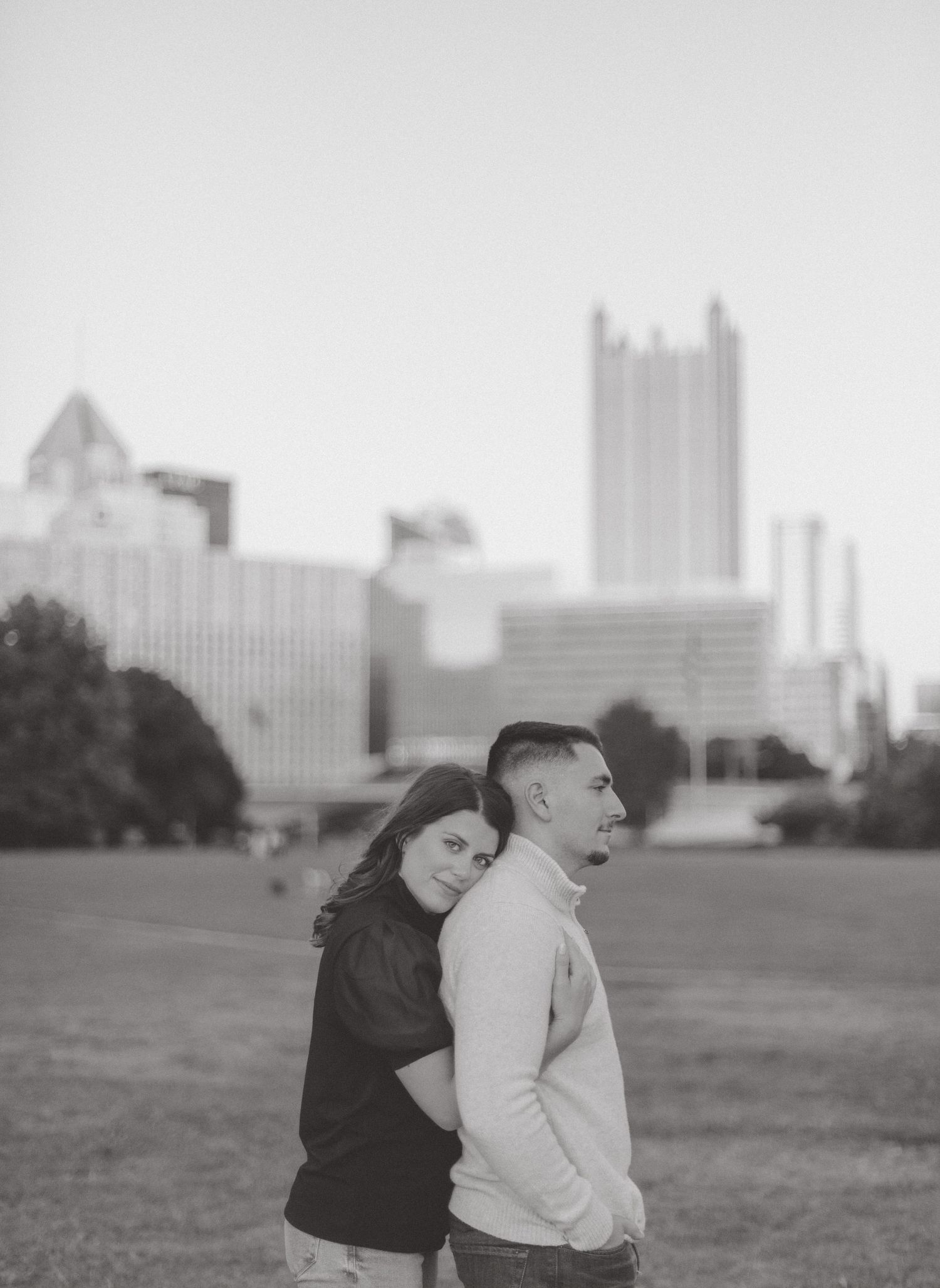 Black and white engagement portrait of a woman hugging her partner from behind on a grassy lawn with a city skyline.